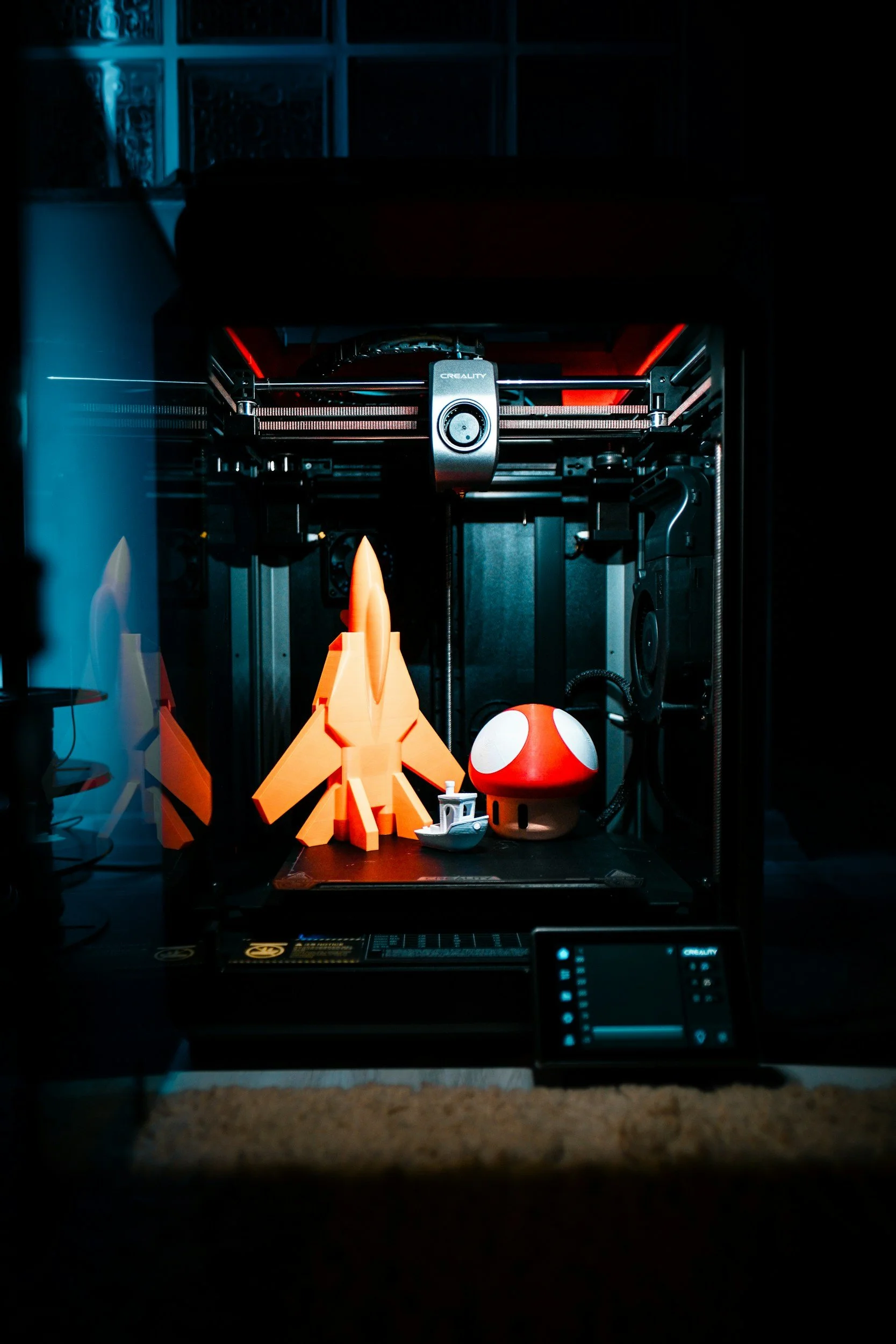 A 3D printer printing a yellow model that resembles a character with a horn, along with a red and white mushroom-shaped object and a tiny floating ship model. The print is inside a dark enclosure with some equipment and a controller visible.