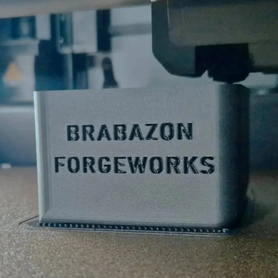 A metal sign with the words "BRABAZON FORGEWORKS" engraved on it, placed on a table or work surface in a workshop or manufacturing setting.
