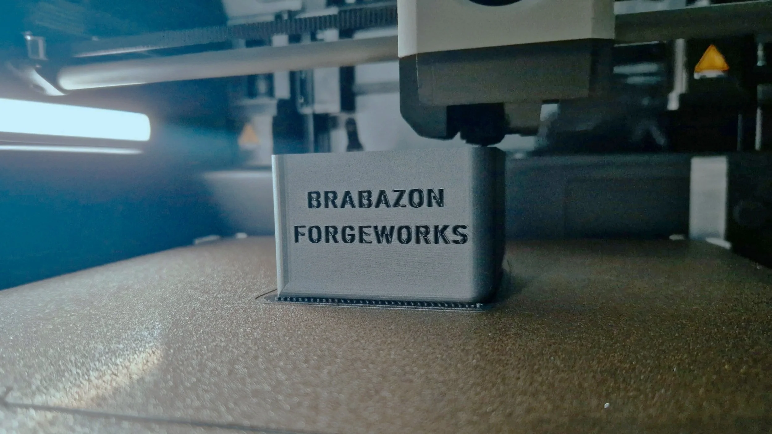 Close-up of a 3D printer in action with a metal component being printed, showing the text 'BRABAZON FORGEWORKS' on the printed piece.