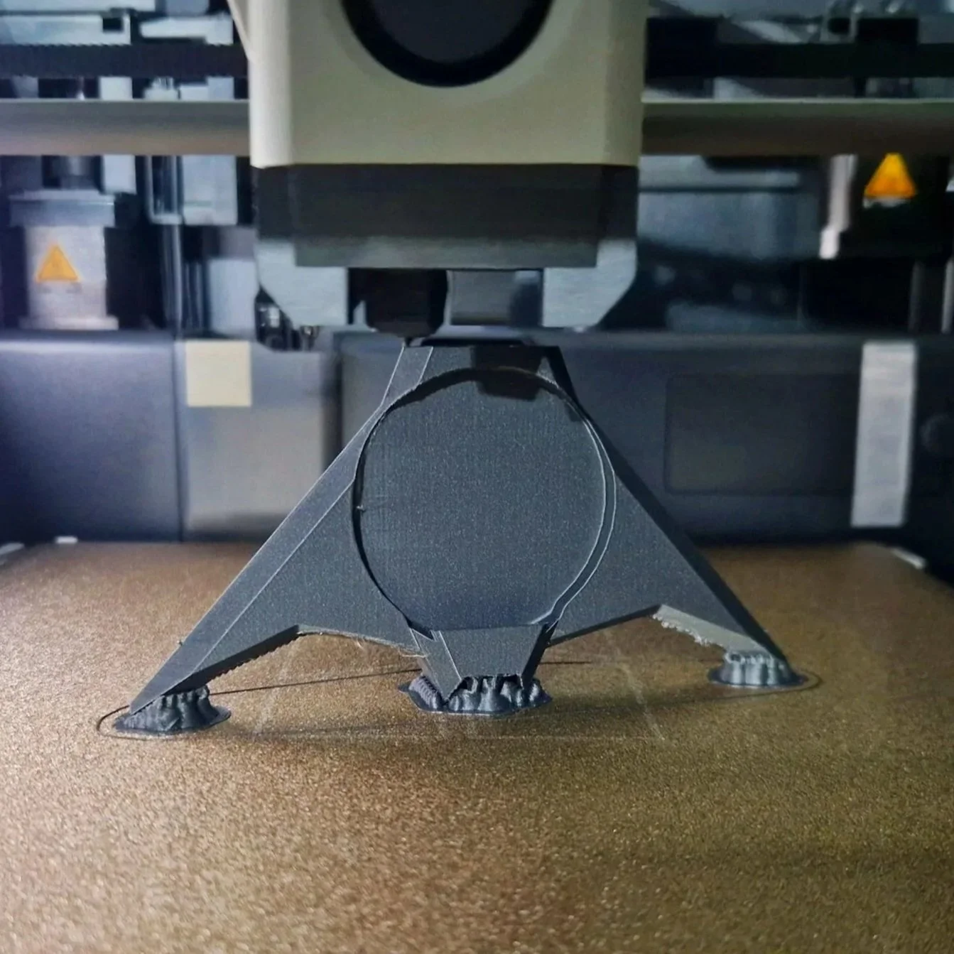 Close-up of a 3D printer's bed with a black filament circle in the center, under the printer's nozzle, with the printer's structural components in the background.