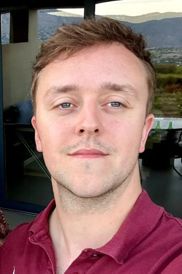 A young man with light skin, short light brown hair, and blue eyes taking a selfie outdoors with mountains and a partly cloudy sky in the background.