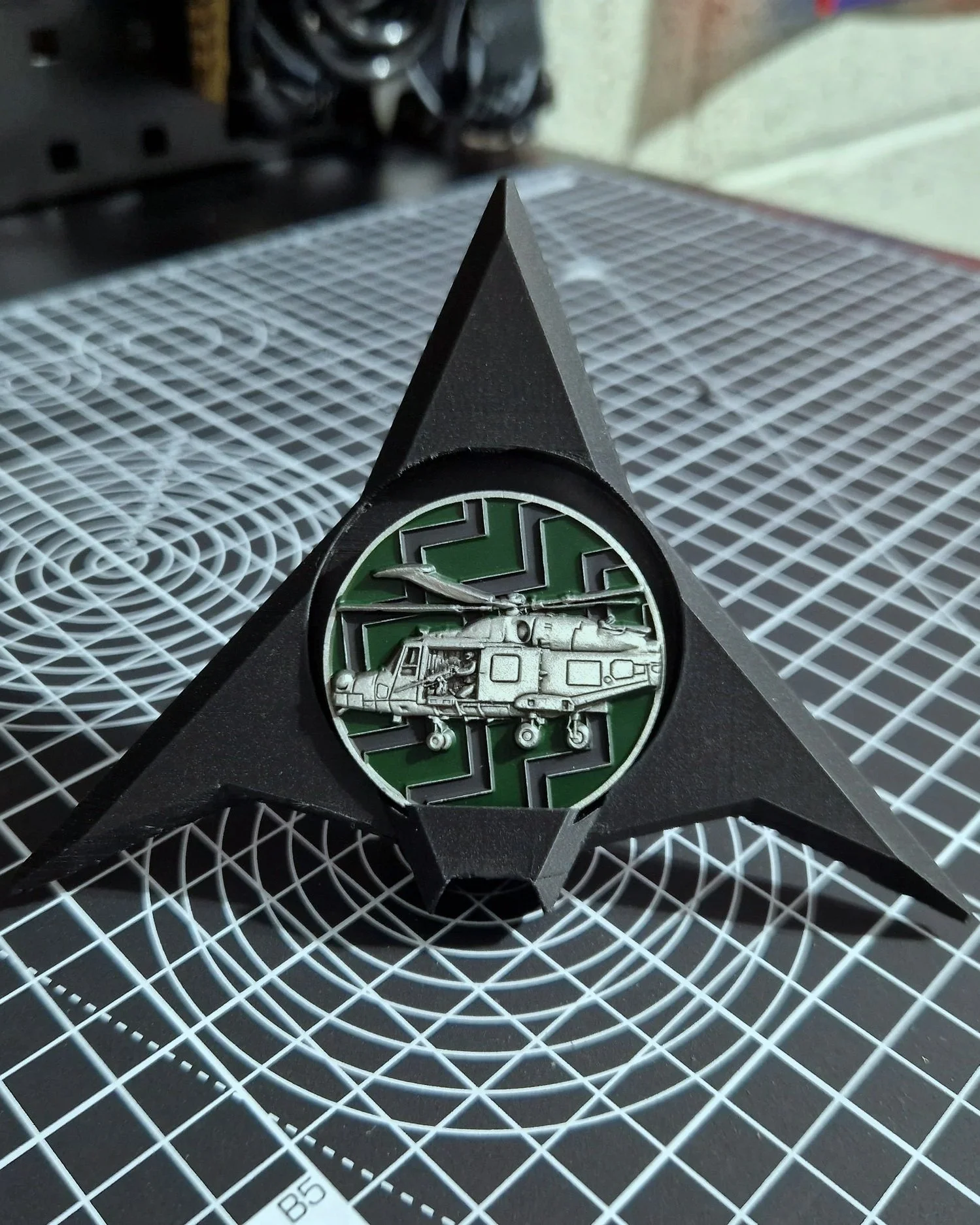 A black triangular object with a metallic logo featuring a tank, mounted at the top of the object. The background contains a gray wire grid and some blurred objects.