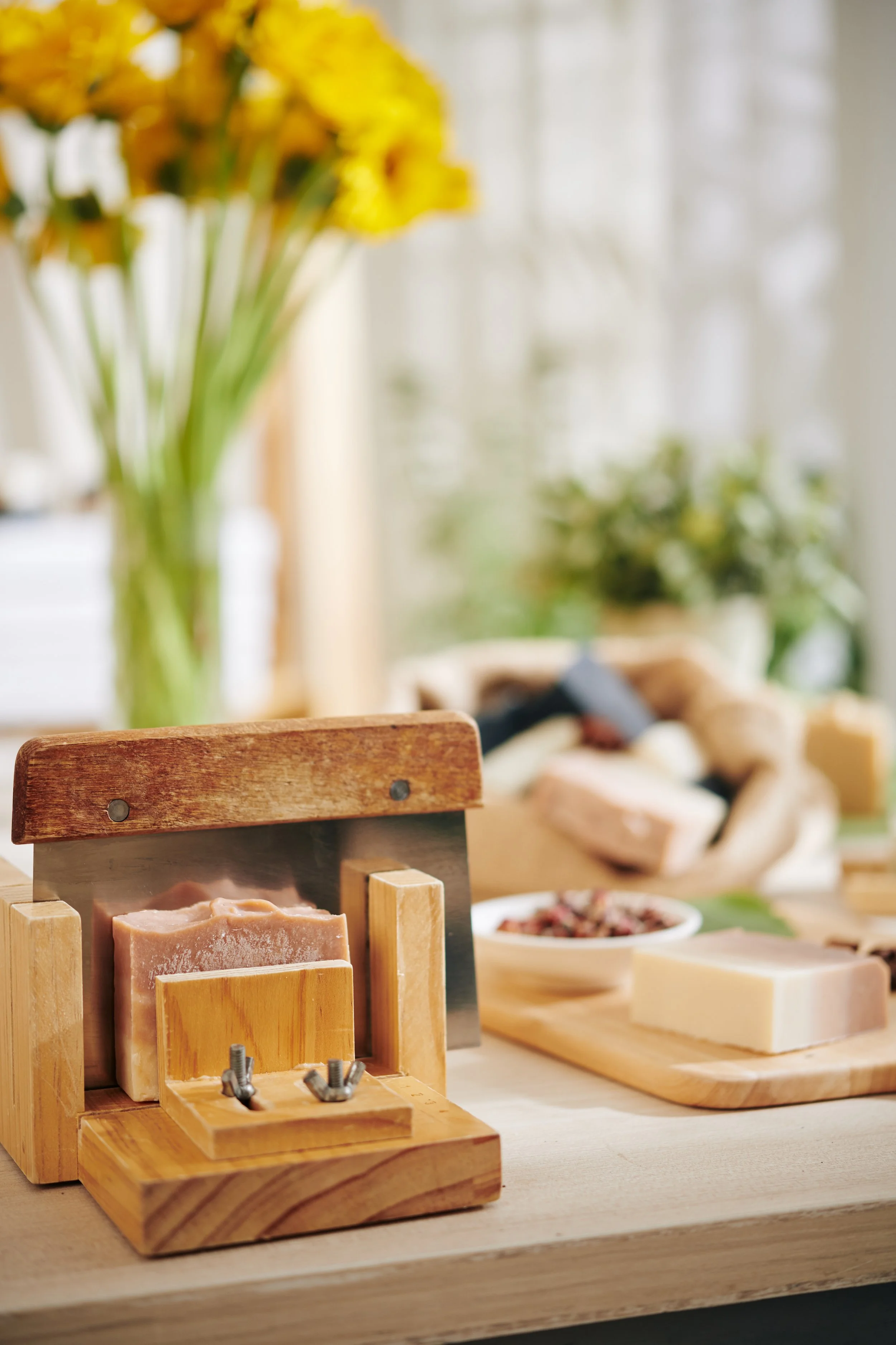 Close-up of soaps in a wooden display rack on a kitchen countertop. In the background, there are flowers, a plate of sliced cheese, and other food items on a wooden cutting board.