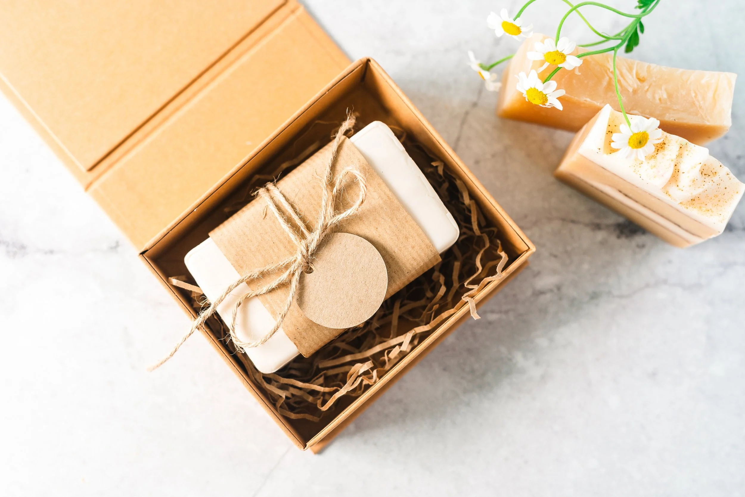 A gift box with a bar of soap wrapped in brown paper and tied with twine, beside two handmade soap bars decorated with daisies and herbs.