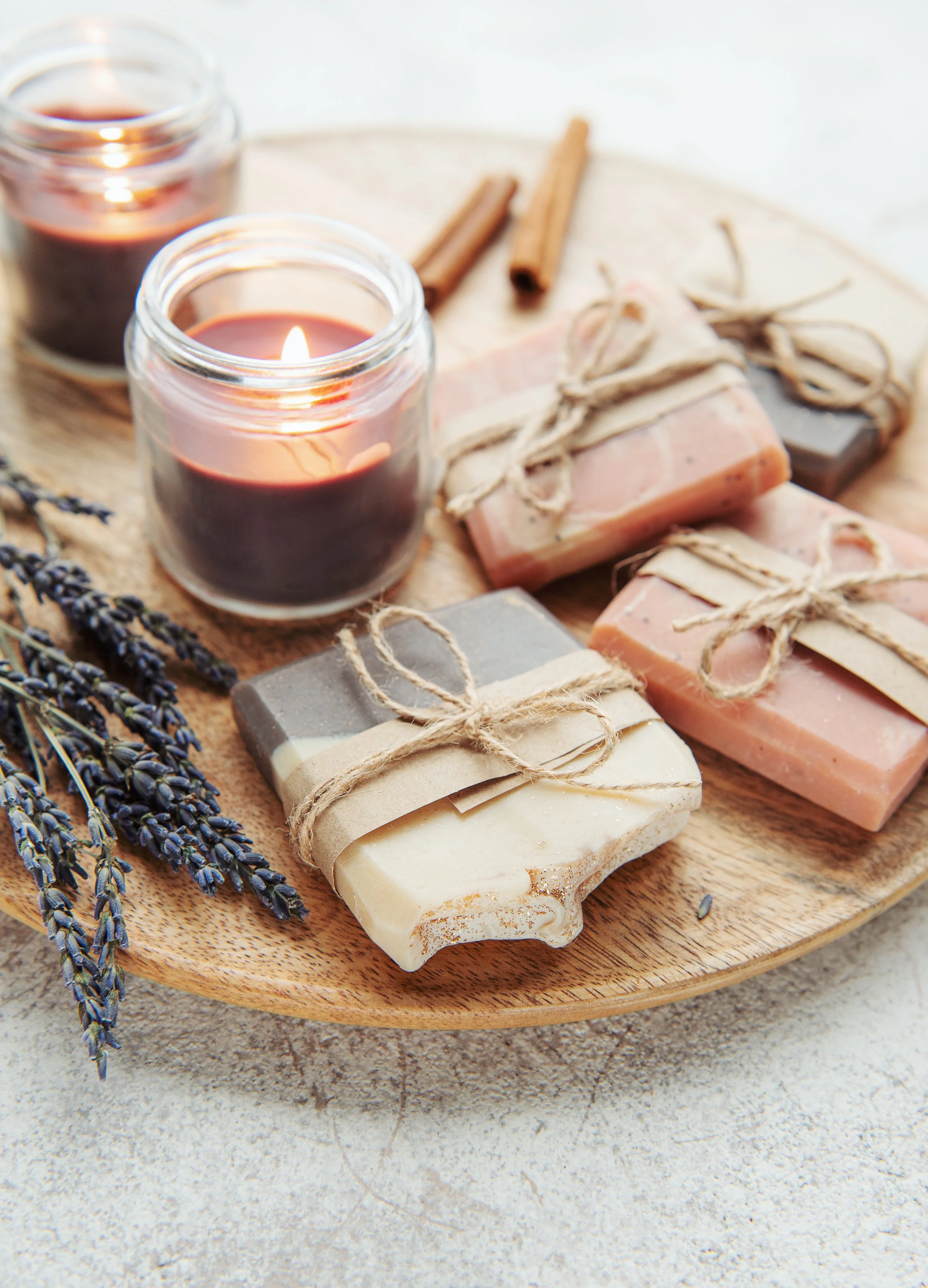 A wooden tray holding lit candles in glass jars, handmade soaps wrapped with twine, and sprigs of lavender lie on a textured surface.