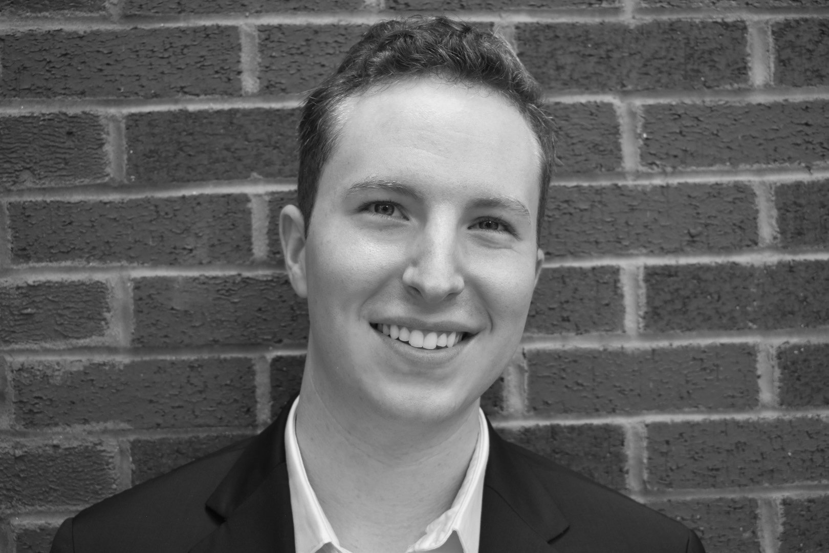 Black and white portrait of a young man with short curly hair, smiling, wearing a suit and a collared shirt, standing in front of a brick wall.