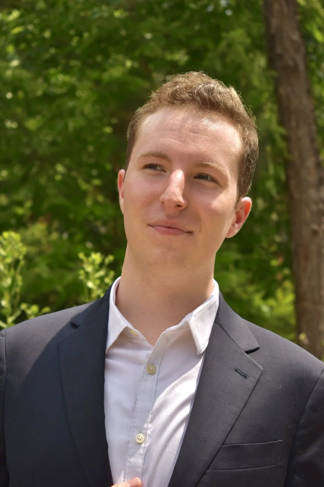 Young man in a suit and white shirt standing outdoors with green trees in the background.