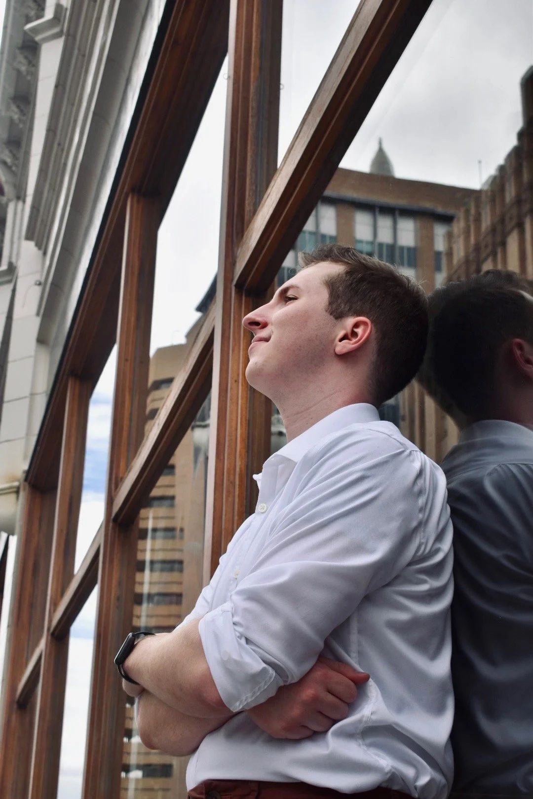 A young man in a white dress shirt with rolled-up sleeves leaning against a wooden window frame, smiling with eyes closed, with a cityscape in the background.