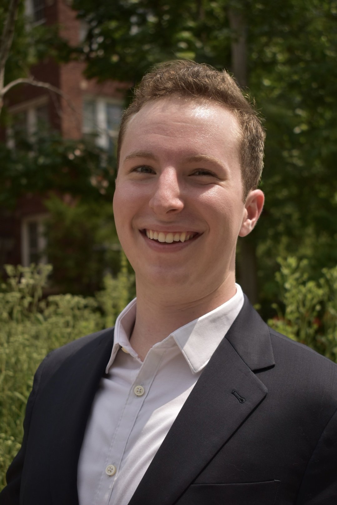 A young man smiling outdoors, wearing a black suit jacket and a white dress shirt, with trees and a building in the background.