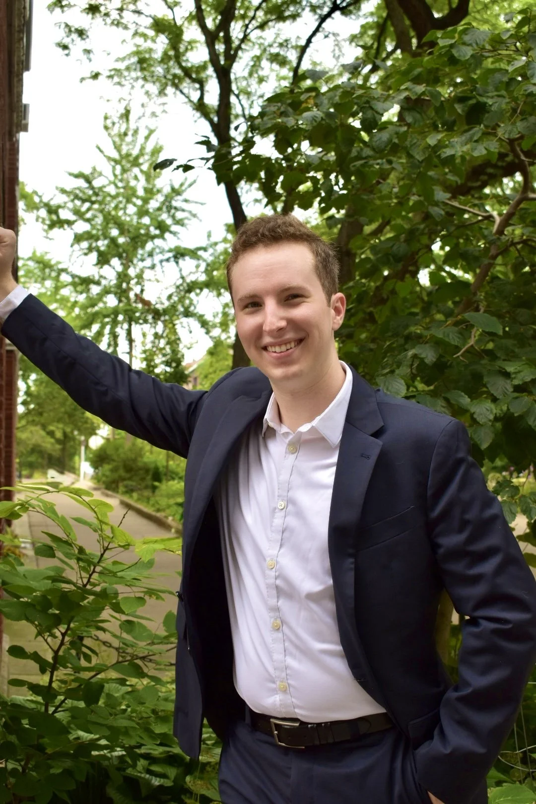 A young man in a navy suit and white shirt smiling outdoors with green trees and bushes in the background.