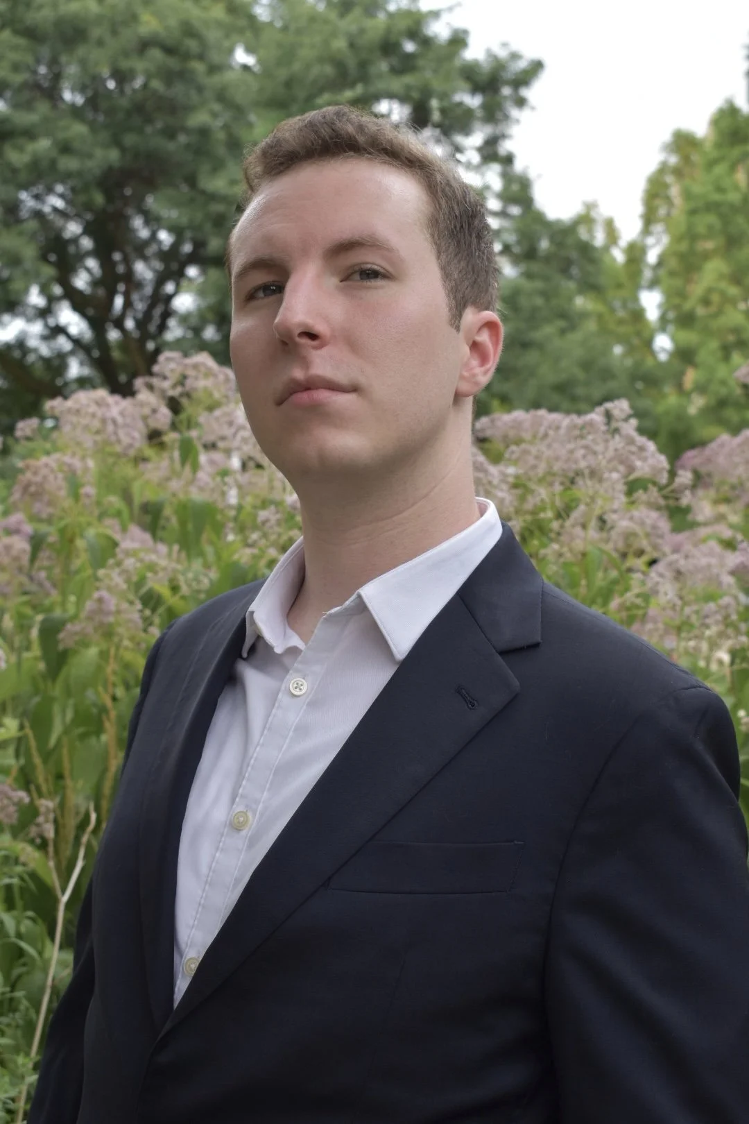 A young man in a black suit and white shirt standing outdoors near flowering plants and trees.
