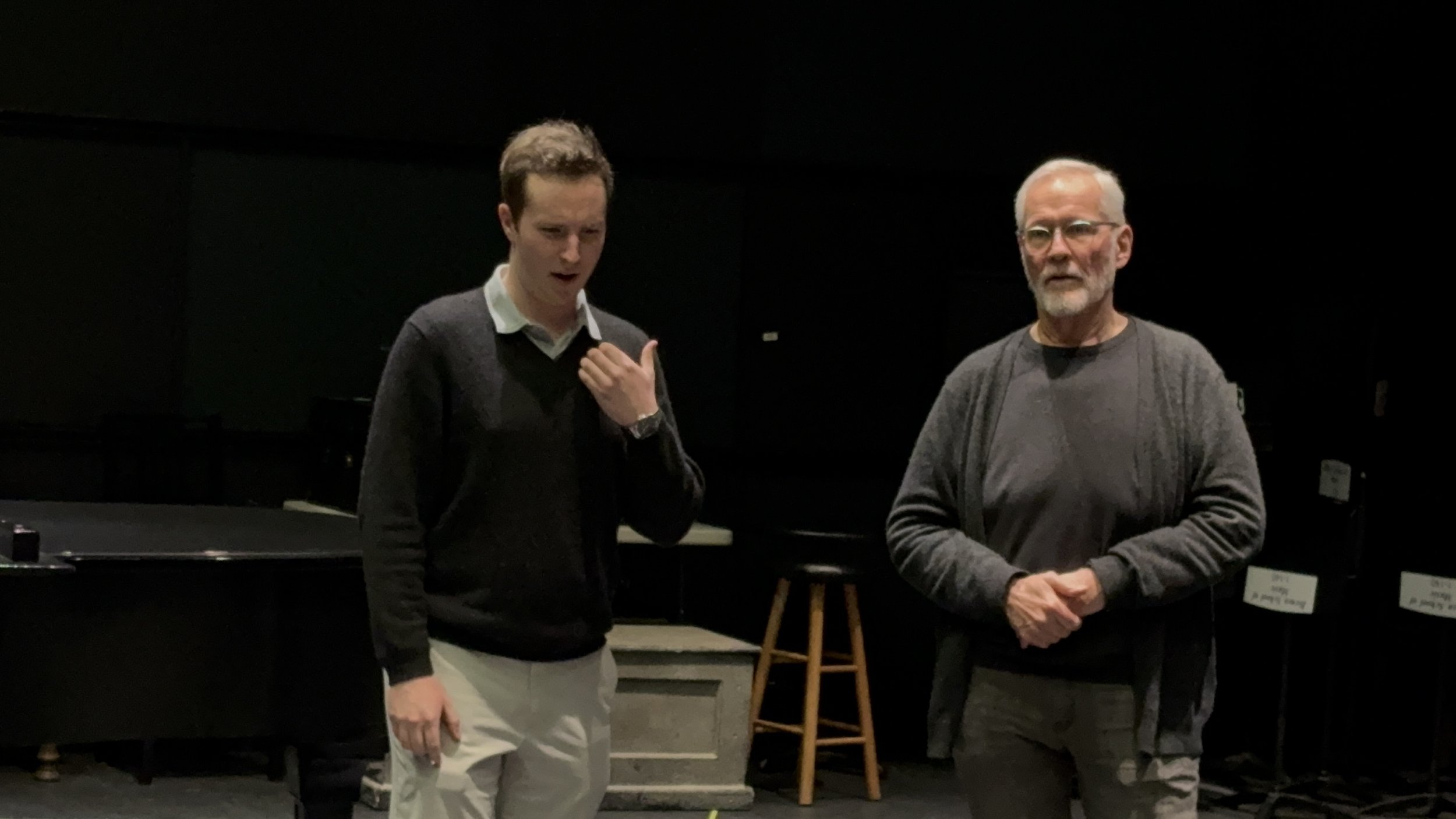 Two men standing in a dark room, one younger with brown hair wearing a dark sweater and light-colored pants, the other older with gray hair, beard, and glasses wearing a gray sweater. They appear to be having a conversation.