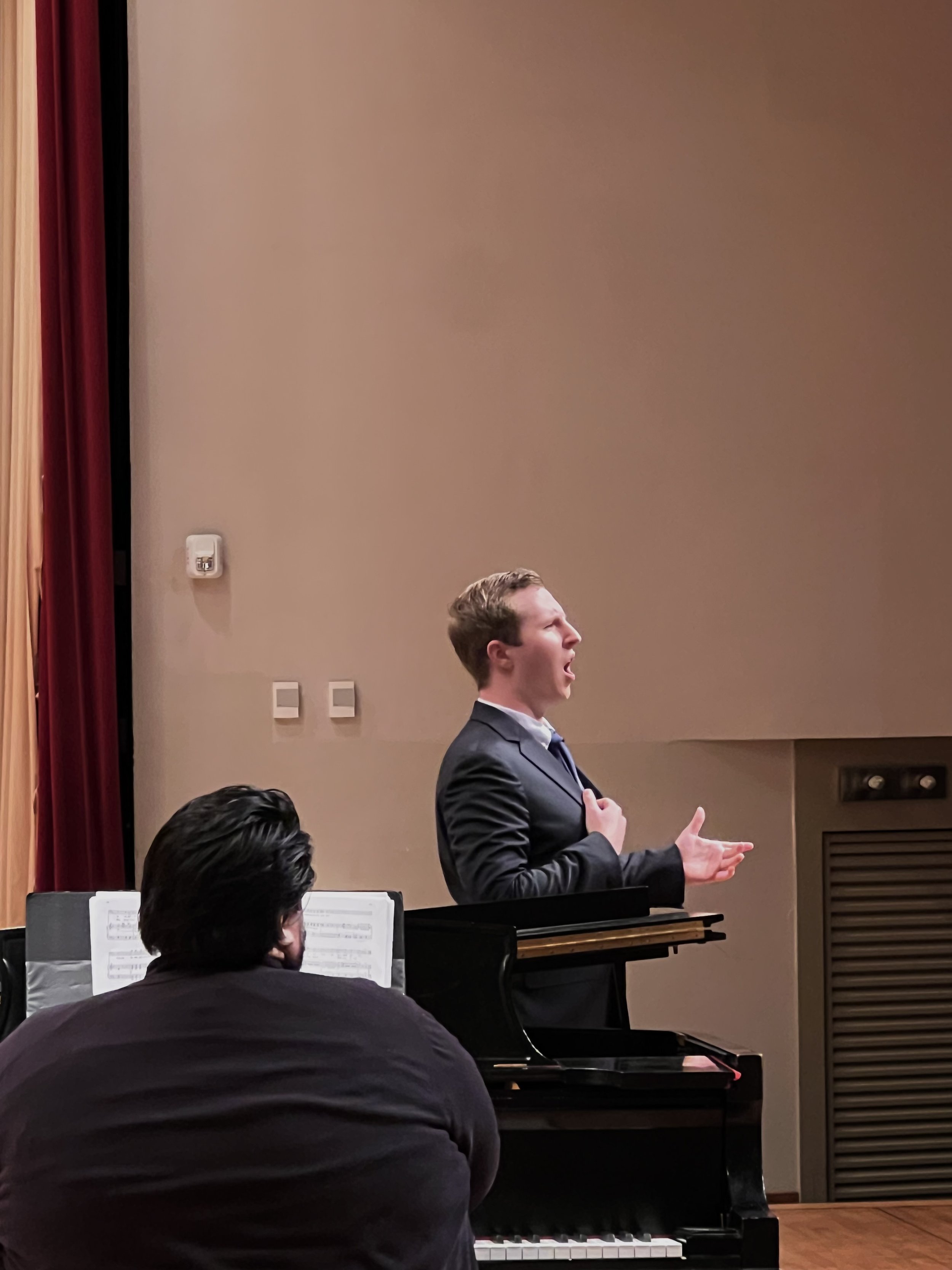 A young man in a suit and blue tie is singing passionately while playing the piano during a performance. There is a pianist seated at the piano in front, with sheet music on top, and part of the audience's head is visible in the foreground. The room has beige walls, red and yellow curtains on the sides, switches and electrical outlets on the wall.