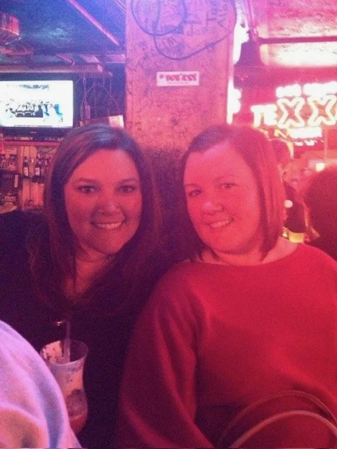 Two women smiling at a bar or restaurant with neon signs and a TV in the background.