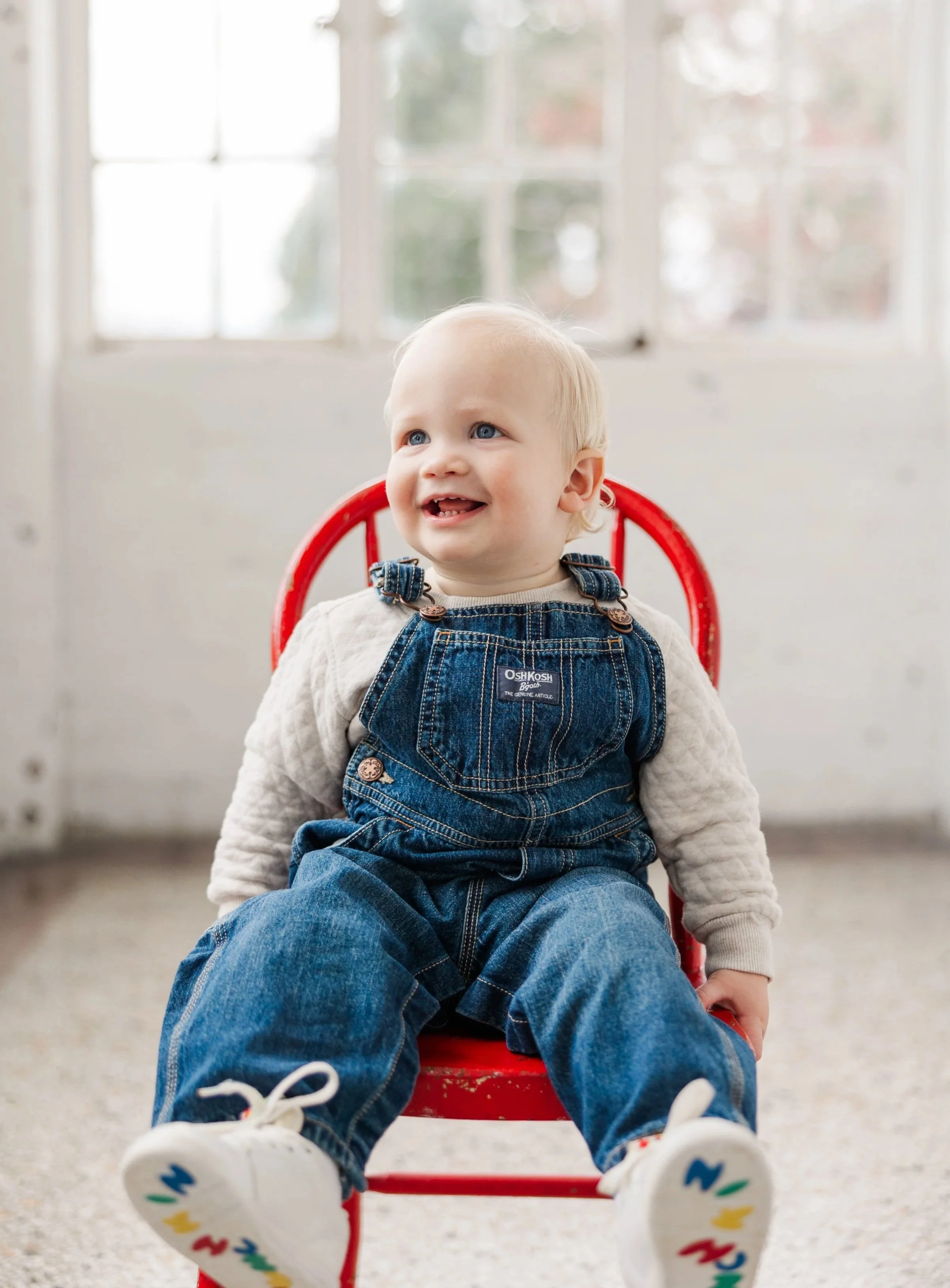 A young child with blonde hair and blue eyes sitting in a red chair, smiling, wearing a beige sweater and denim overalls, with white sneakers featuring colorful lightning bolt designs, in front of a white wall and windows with blurred outdoor scenery.