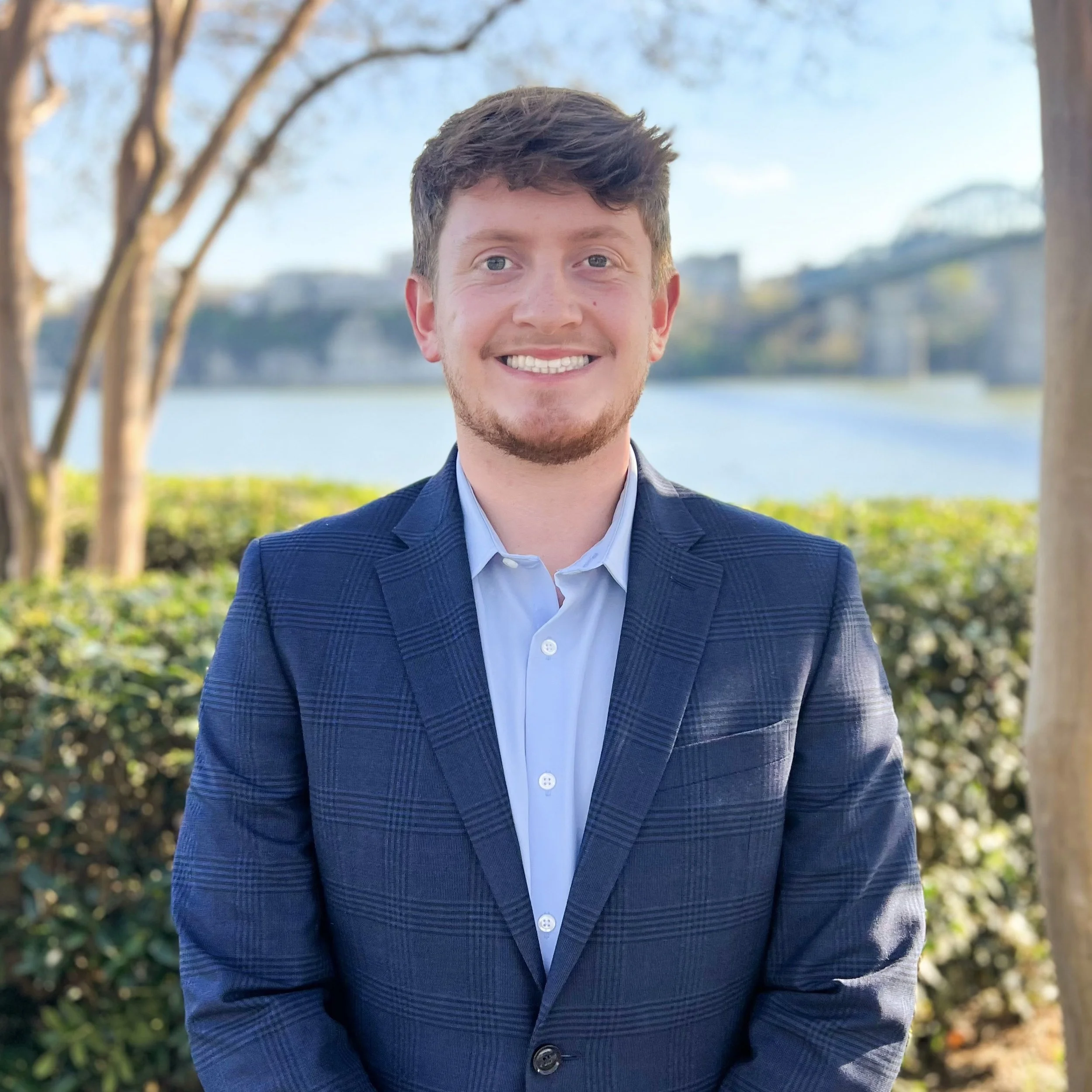 A young man with brown hair and a beard smiling outdoors near a body of water and trees, dressed in a blue checkered blazer and light blue shirt.