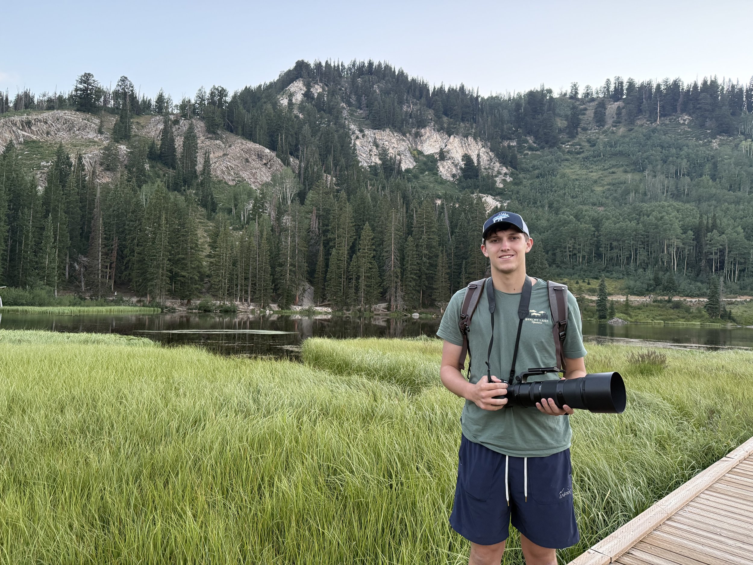 A young man standing in a grassy field holding a pair of binoculars, with a scenic backdrop of a forested mountain and a lake.