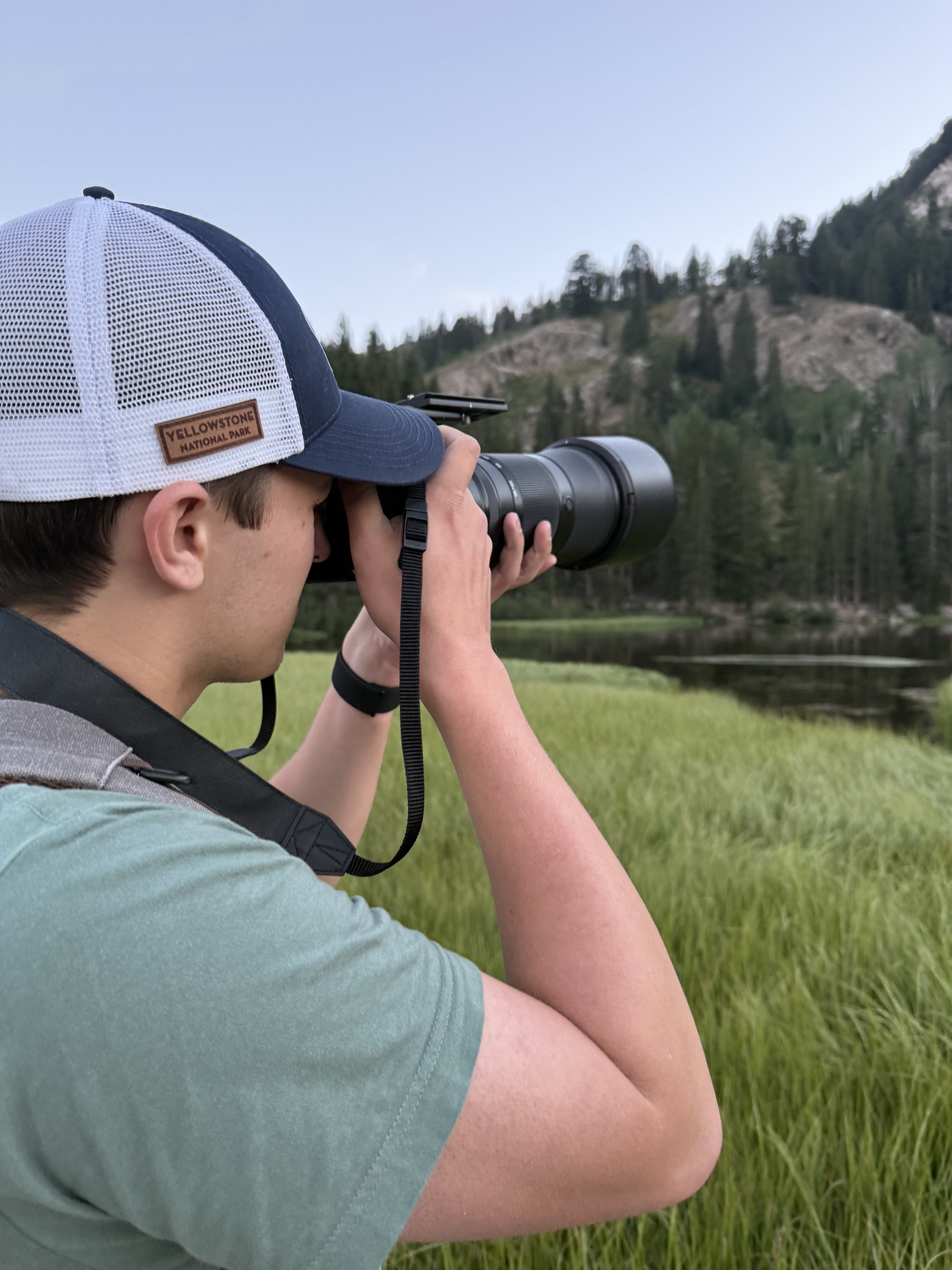 A person wearing a gray t-shirt and a white and navy blue trucker hat with a "Yellowstone National Park" label, holding a camera with a long lens and taking a photograph of the mountain and woods in the background.