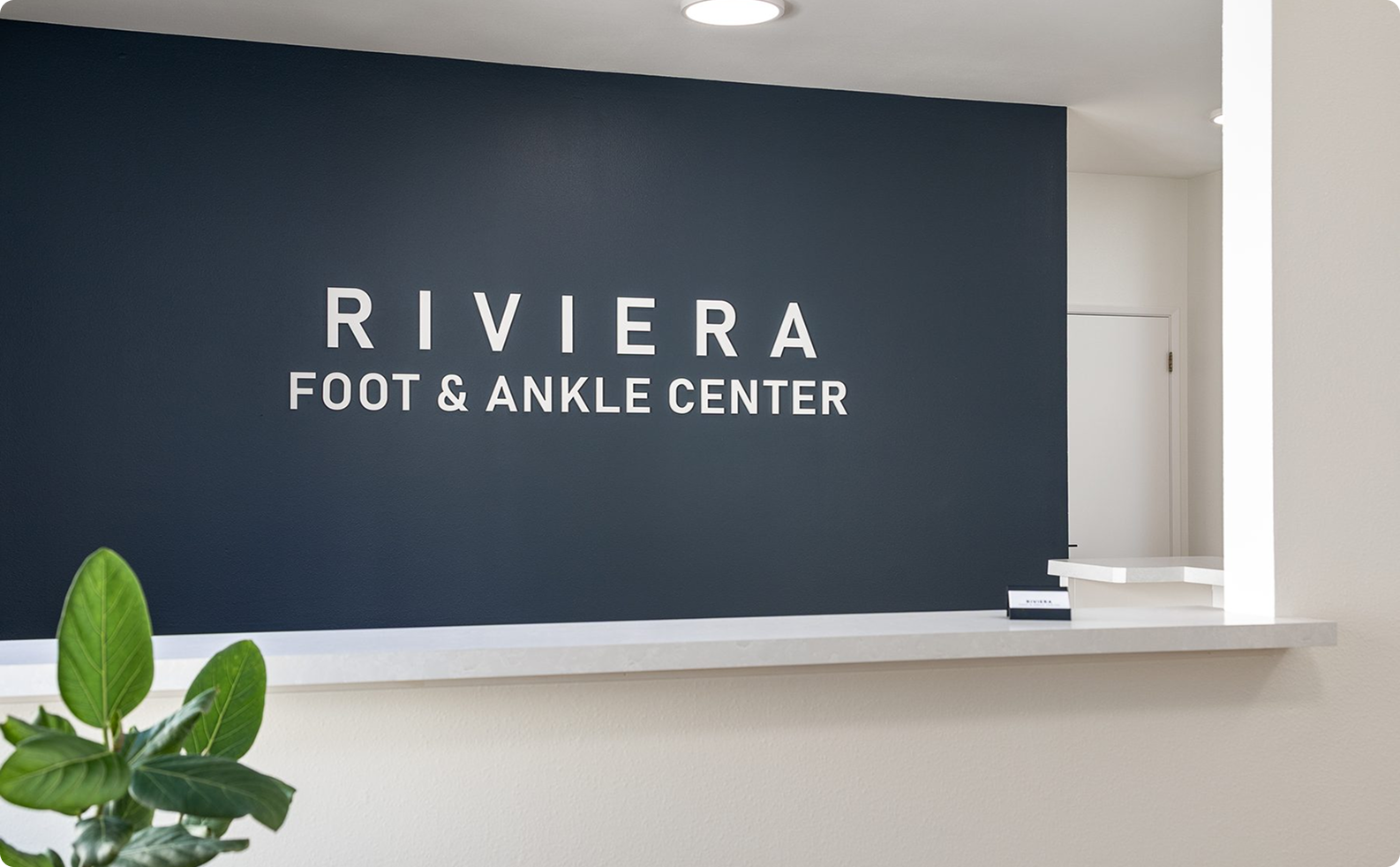 Reception area of Riviera Foot & Ankle Center with a dark blue accent wall and white signage, beige counter, and a potted green plant in the foreground.