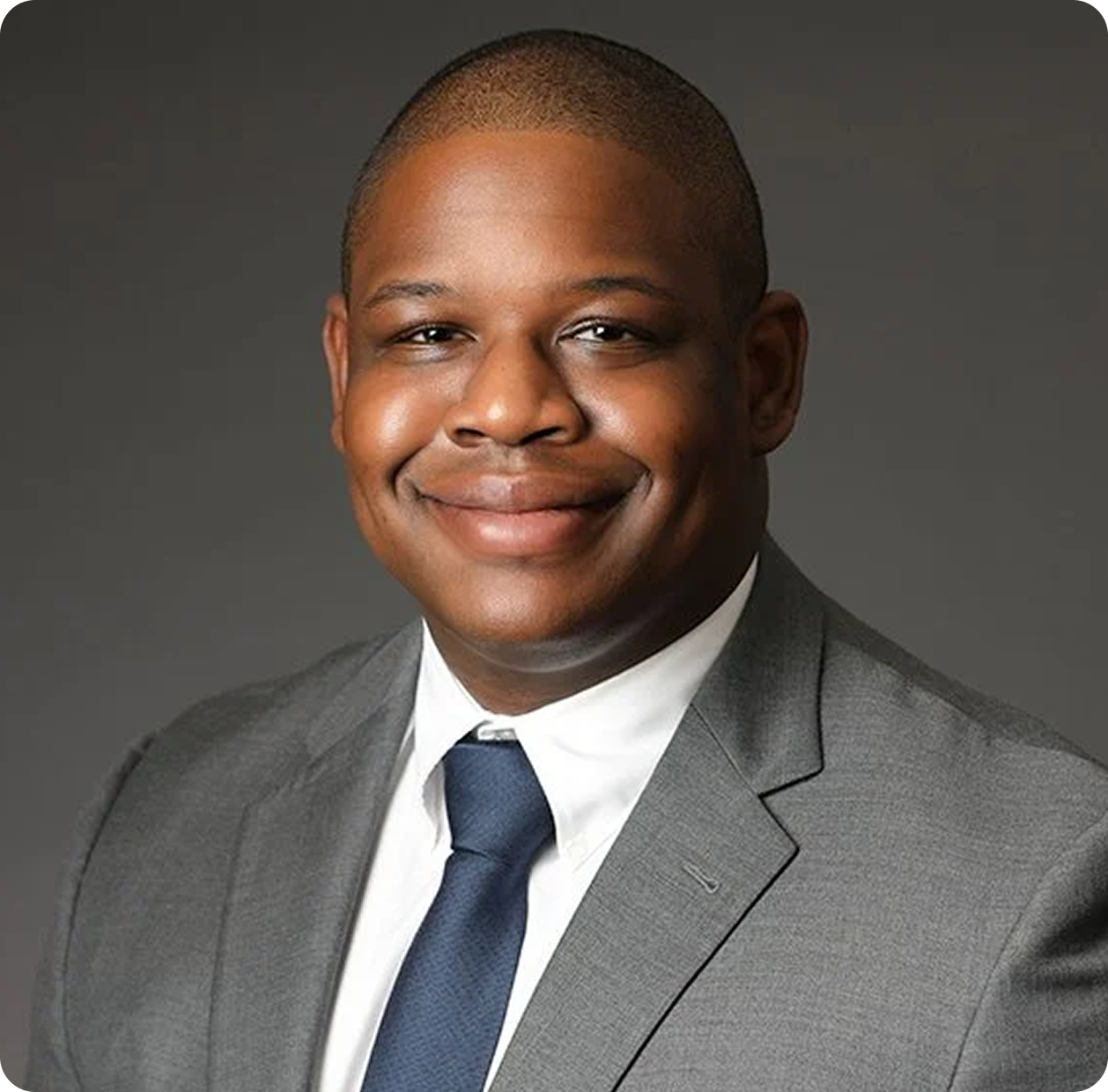 Professional headshot of a smiling African American man wearing a gray suit, white shirt, and blue tie, against a dark gray background.