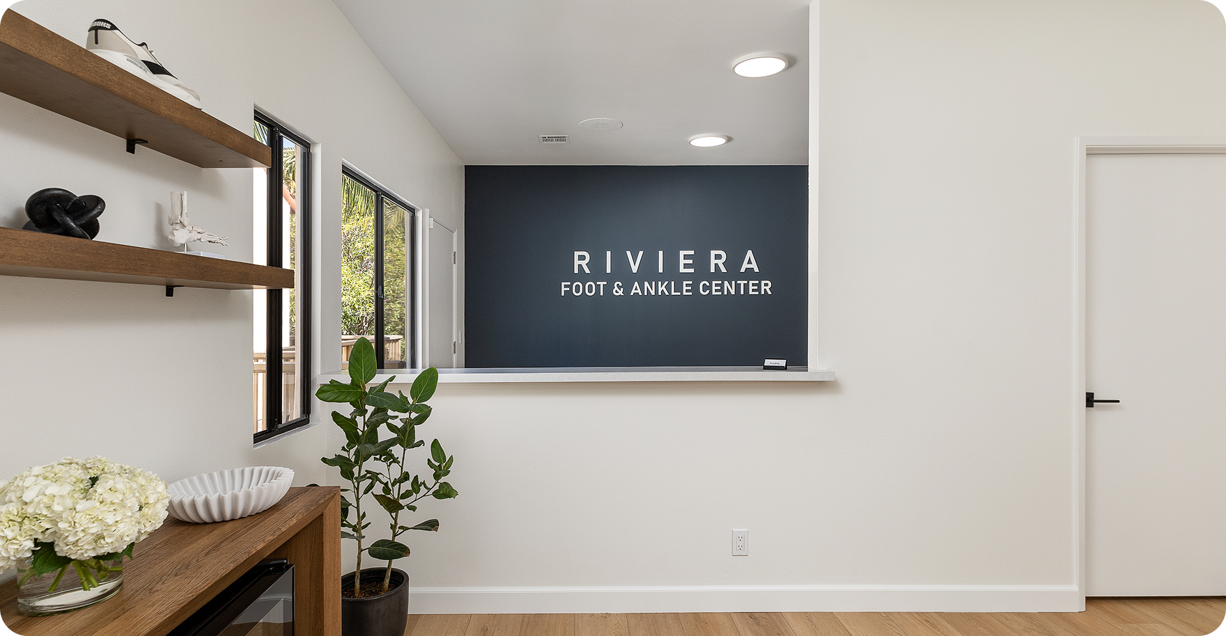 Reception area of Riviera Foot & Ankle Center with a dark blue wall, white sign, wooden shelves with shoes and decorative objects, a potted plant, and a white countertop.