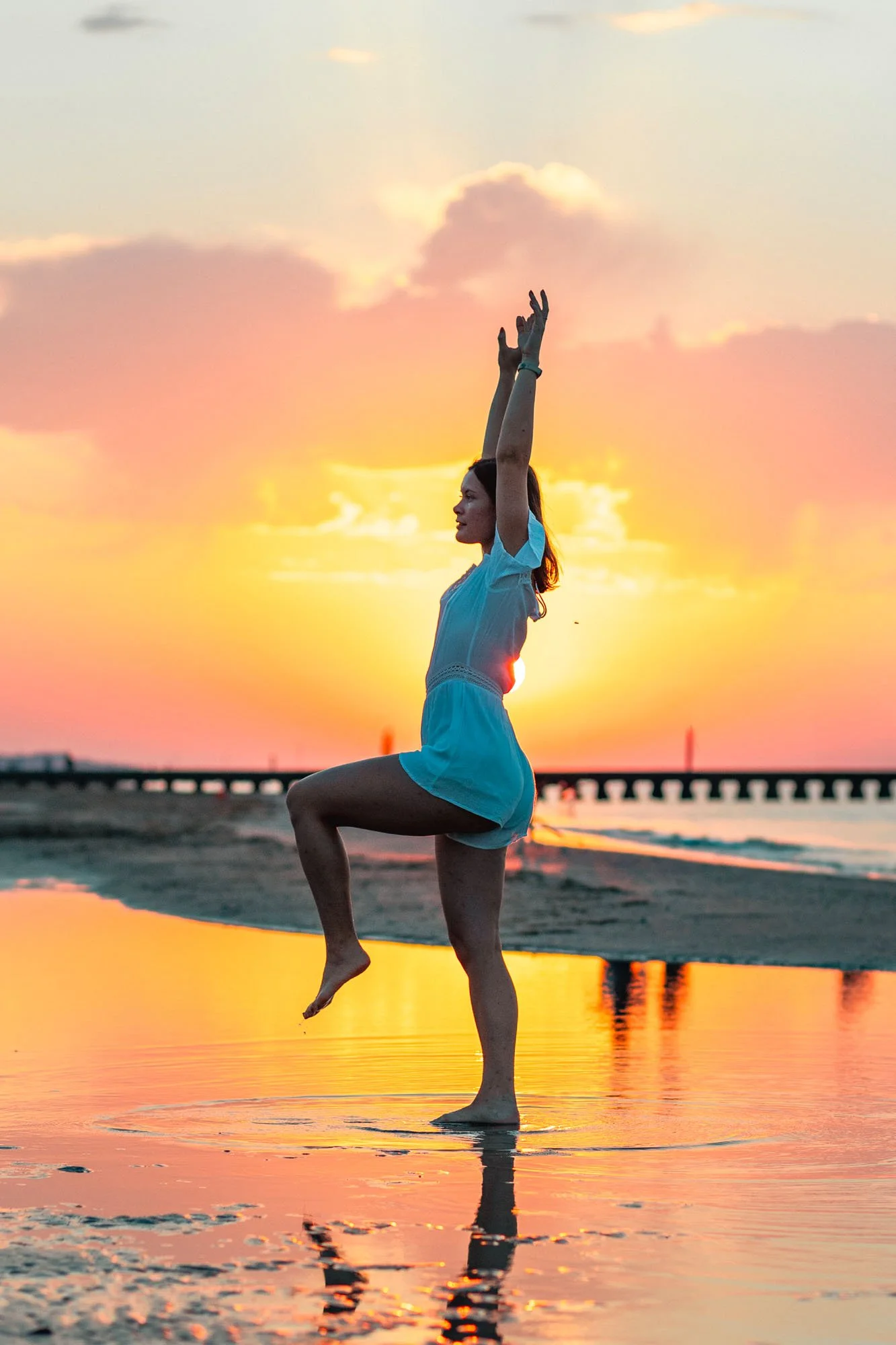 Una donna in posizione di yoga sul mare al tramonto, con il cielo colorato di rosa e arancione.