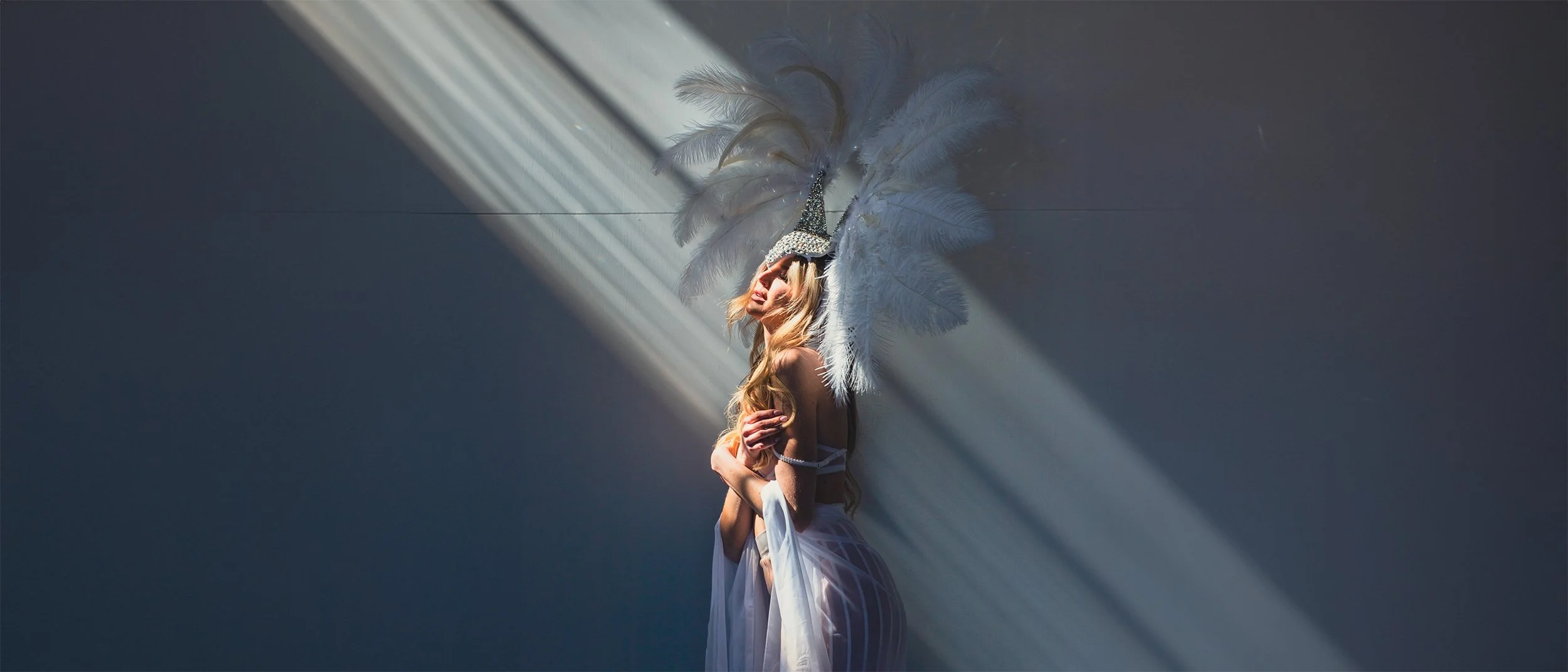 Donna con un costume da carnevale con grandi piume bianche che indossa un cappello decorato con cristalli, seduta contro il muro, illuminata da una luce diagonale.