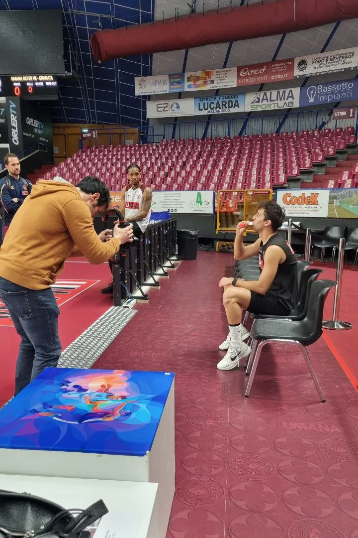 Due giocatori di pallacanestro seduti su sedie in un palazzetto dello sport mentre uno di loro mangia uno snack, mentre un uomo con una macchina fotografica li sta fotografando, altri persone sono sullo sfondo e si vedono le gradinate vuote.