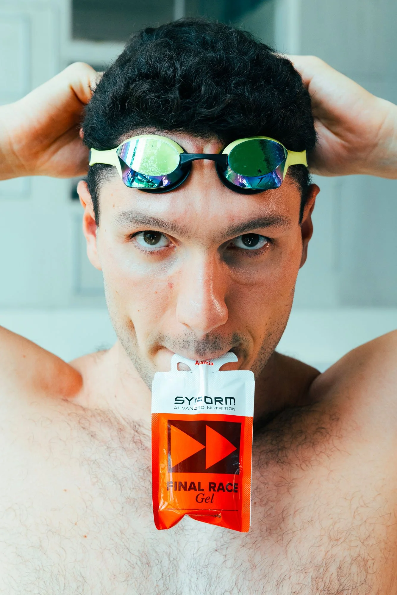 Uomo con occhialini da nuoto sopra la fronte, tiene le mani dietro la testa, e inghiotte un gel energetico durante una competizione di triathlon o corsa.