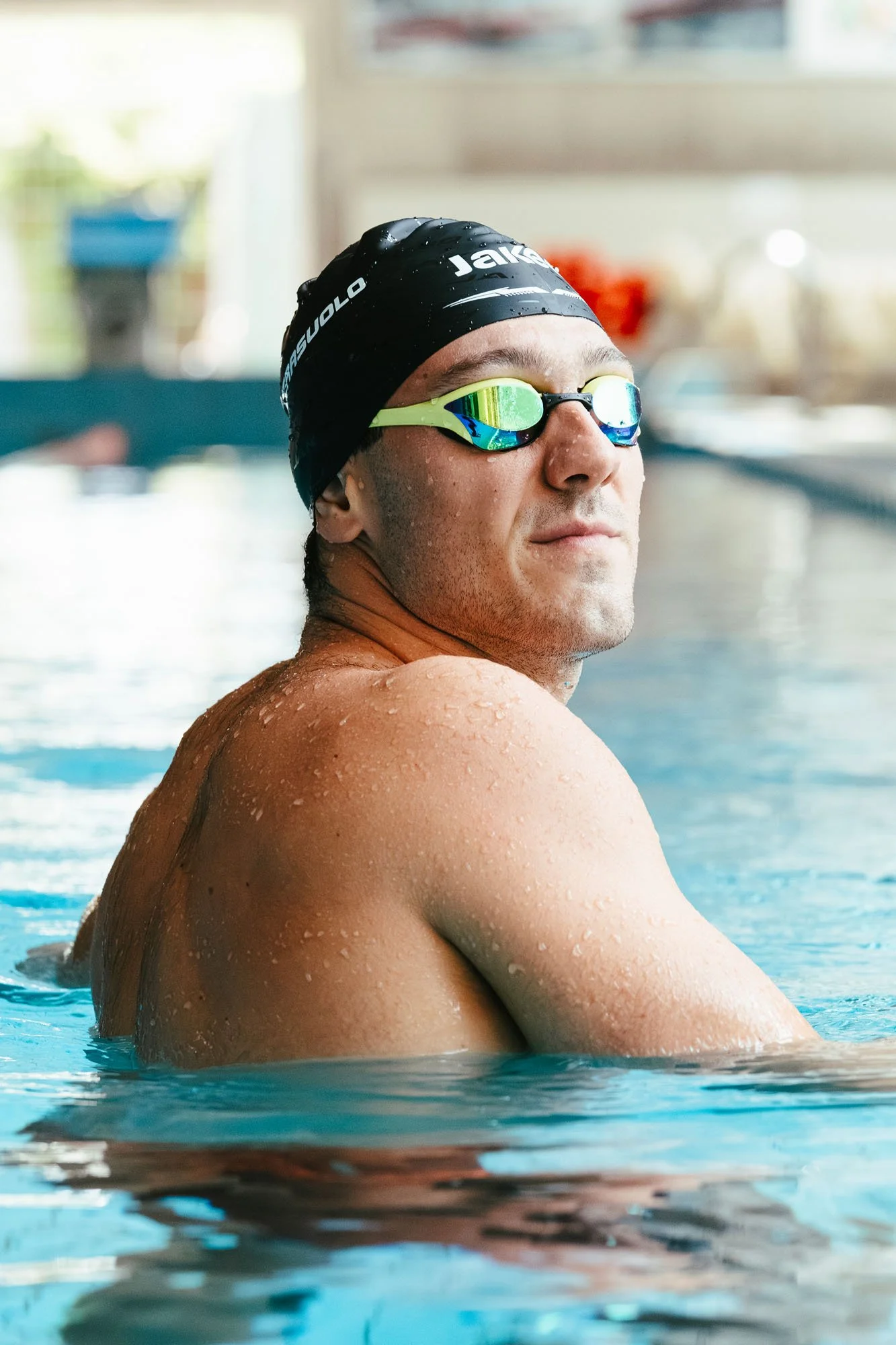 Nuotatore maschio in corsia di piscina, indossa cuffia nera e occhialetti da nuoto con lenti verdi riflettenti, capelli bagnati, con espressione serena.