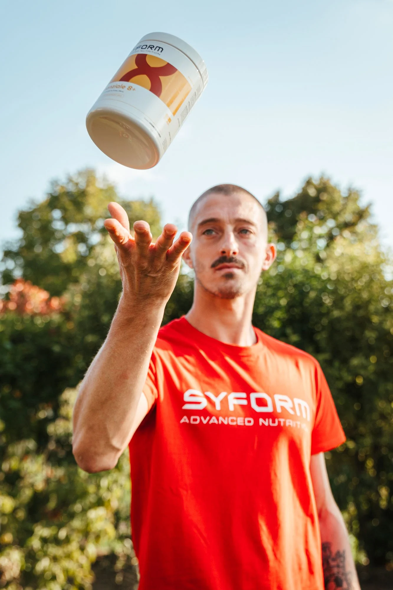 Uomo con maglietta rossa che lancia una bottiglia di integratore alimentare Syform Advanced Nutrition in aria in un parco durante il giorno.