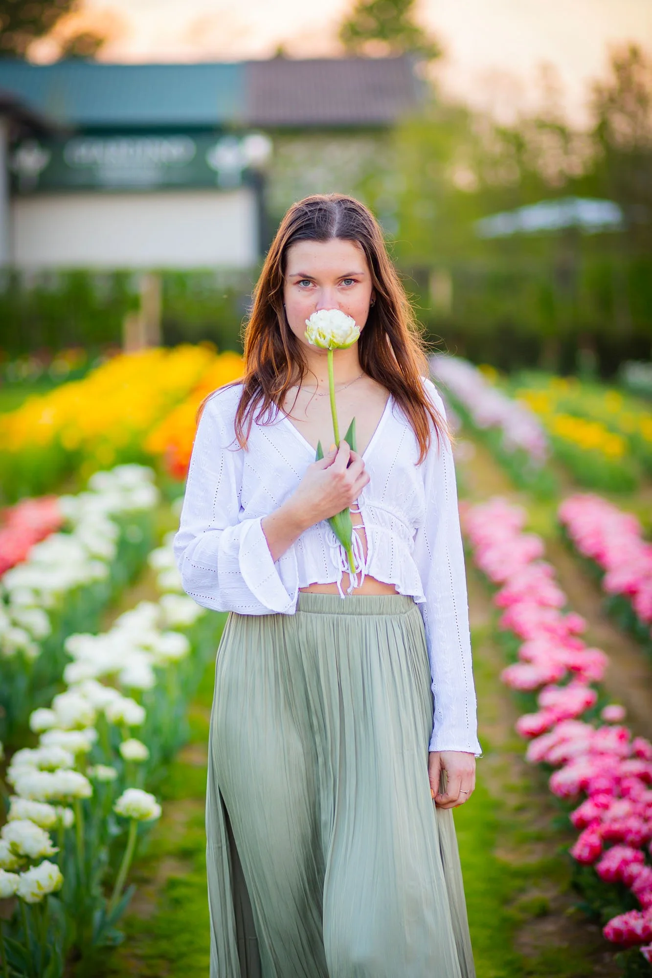 Una giovane donna con capelli castani, indossa un top bianco e una gonna lunga color panna, si trova in un campo di fiori colorati, tenendo un fiore bianco di fronte alla bocca.