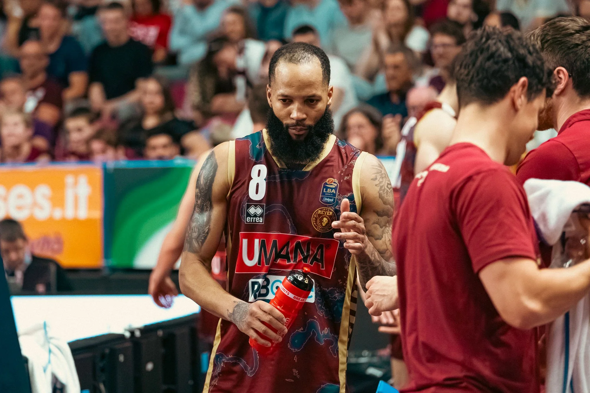 Calciatore con barba e tatuaggi che tiene una borraccia rossa durante una partita, con pubblico sullo sfondo.