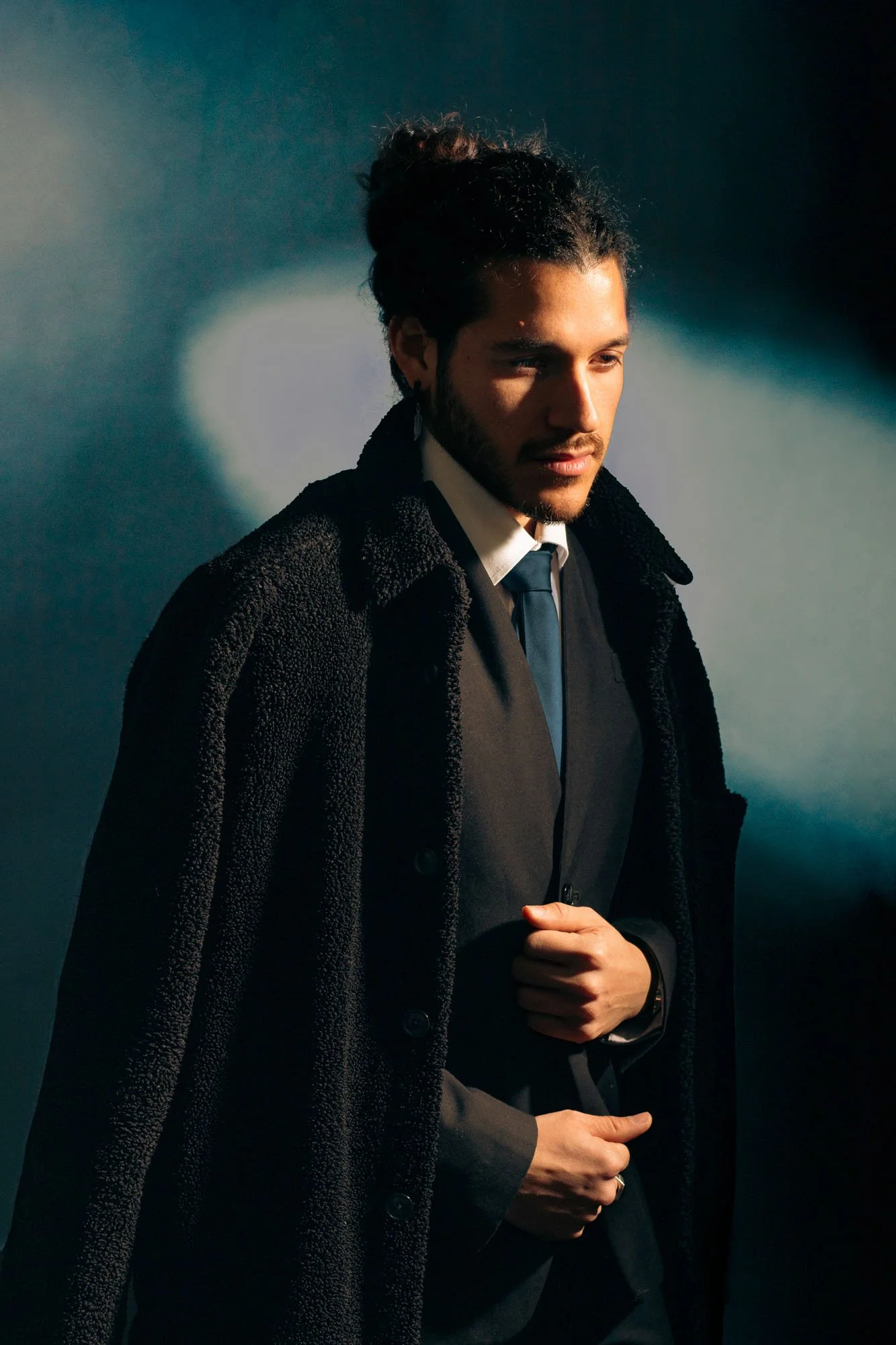 Ritratto di un uomo con barba, capelli raccolti, abbigliamento elegante, posa pensierosa, sfondo scuro con sfumature di luce