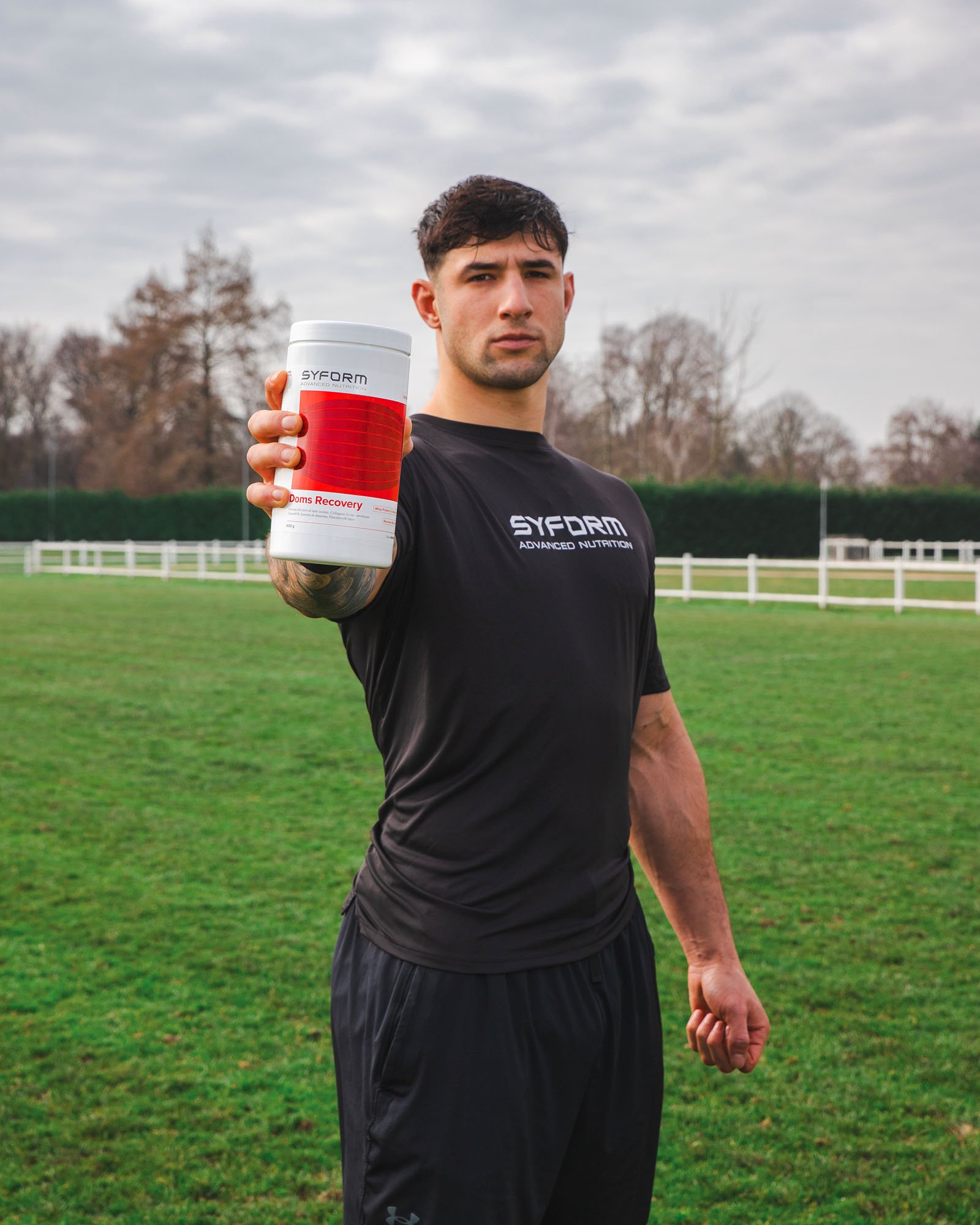 Uomo in tenuta sportiva nera che tiene una scatola di integratori chiamata SYFORM Doms Recovery, in un campo di calcio all'aperto con cielo nuvoloso.