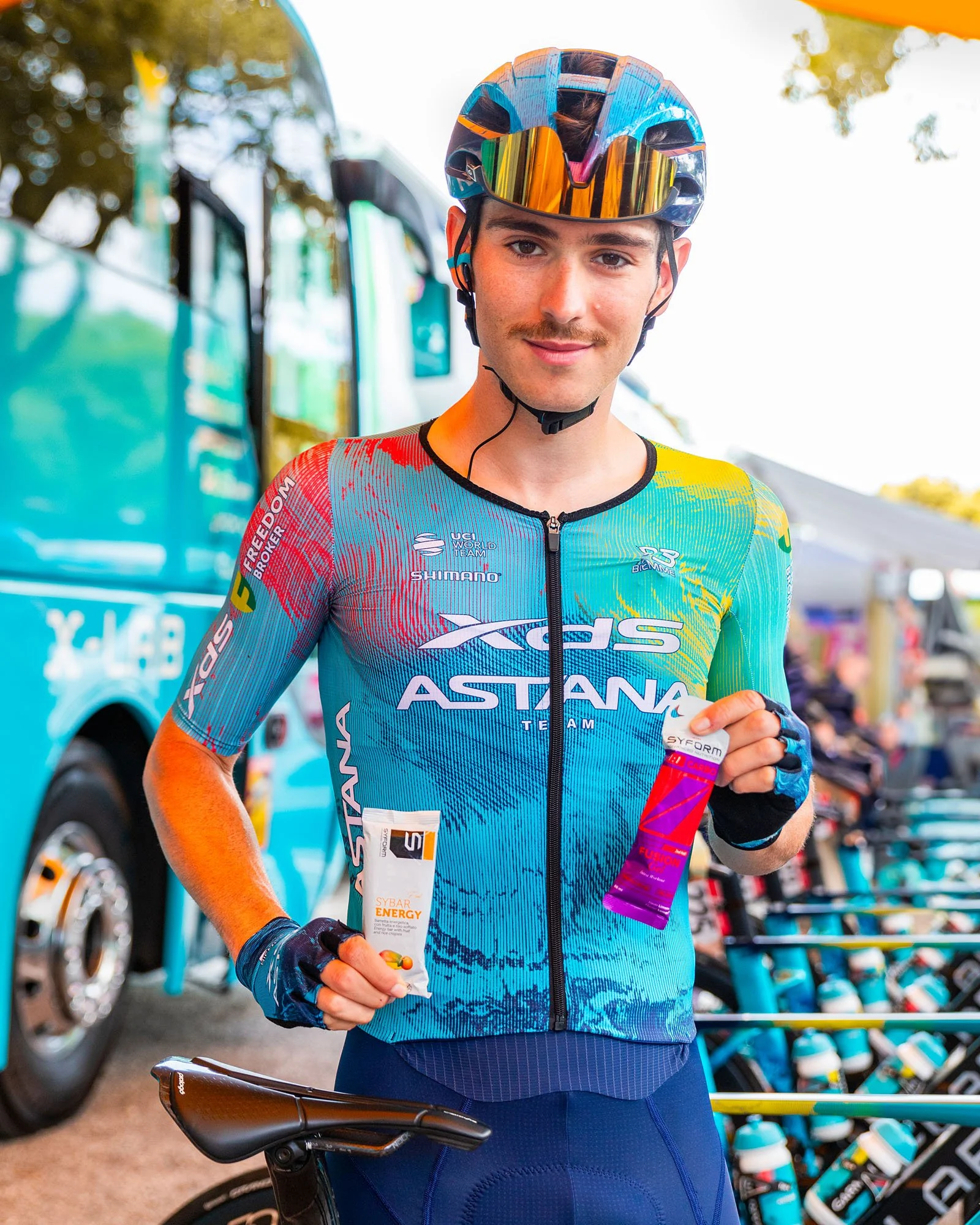 Un ciclista maschio sorridente con casco e abbigliamento sportivo, che tiene una barretta energetica in mano, davanti a un bus e a biciclette di club ciclistici.