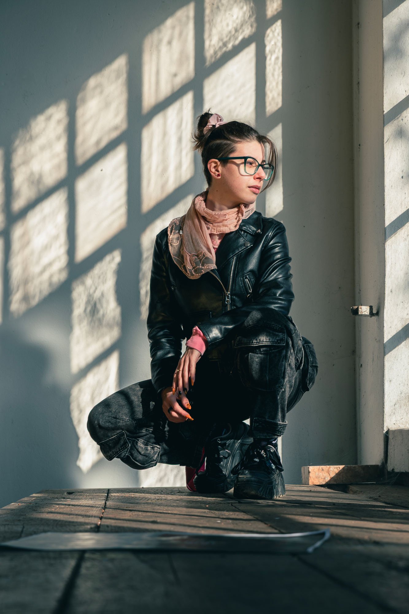 Ragazza con capelli raccolti, indossa occhiali, giacca di pelle, sciarpa rosa e jeans neri, seduta in un ambiente con luce naturale e ombre proiettate da una finestra.