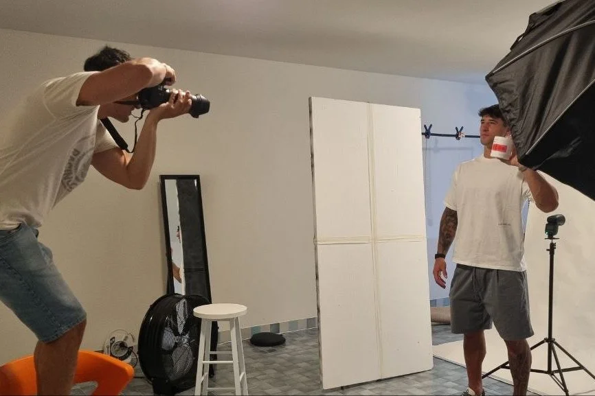 Un uomo fa una sessione fotografica in casa, mentre un altro scatta una foto, posando con un bicchiere in mano e un grande softbox davanti a lui.