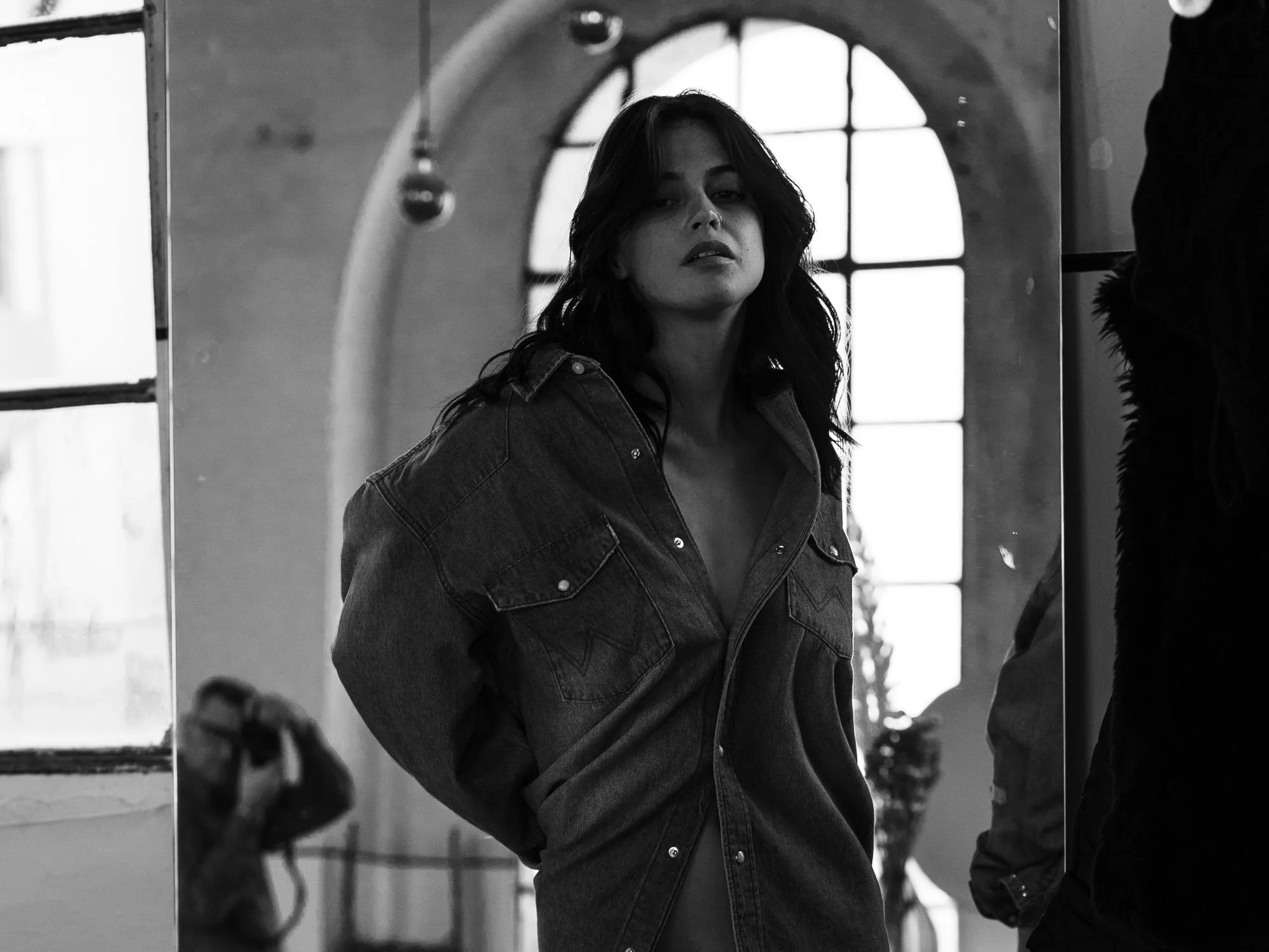 Ritratto in bianco e nero di una donna con i capelli sciolti, indossa una giacca di jeans e guarda verso l'obiettivo, con finestra ad arco sullo sfondo.