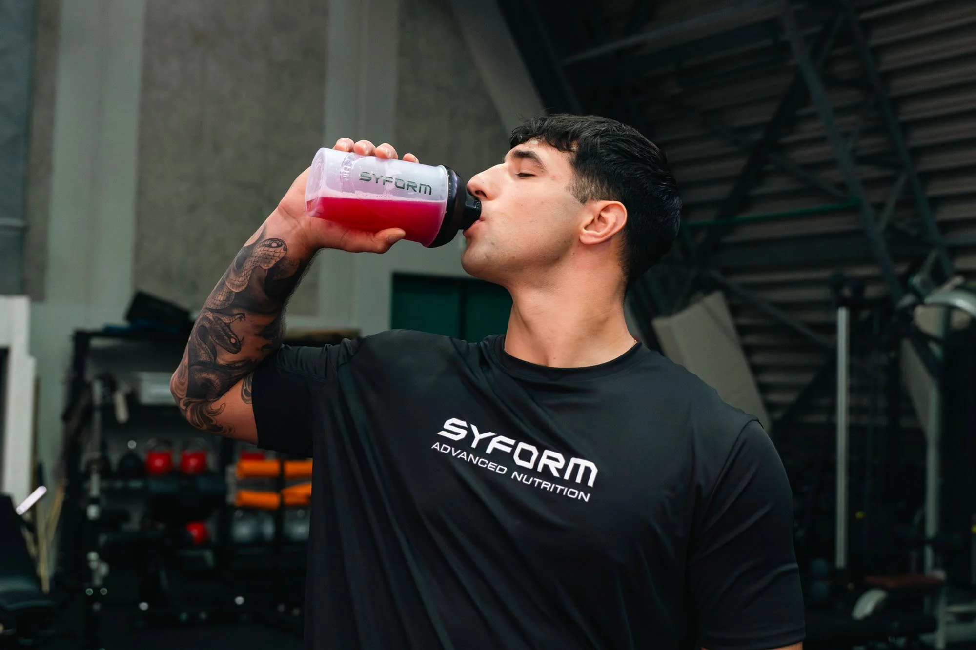 Un uomo muscoloso con tatuaggi sul braccio destro beve da una borraccia rosa in un ambiente di palestra. Indossa una maglietta nera con il logo Syform.