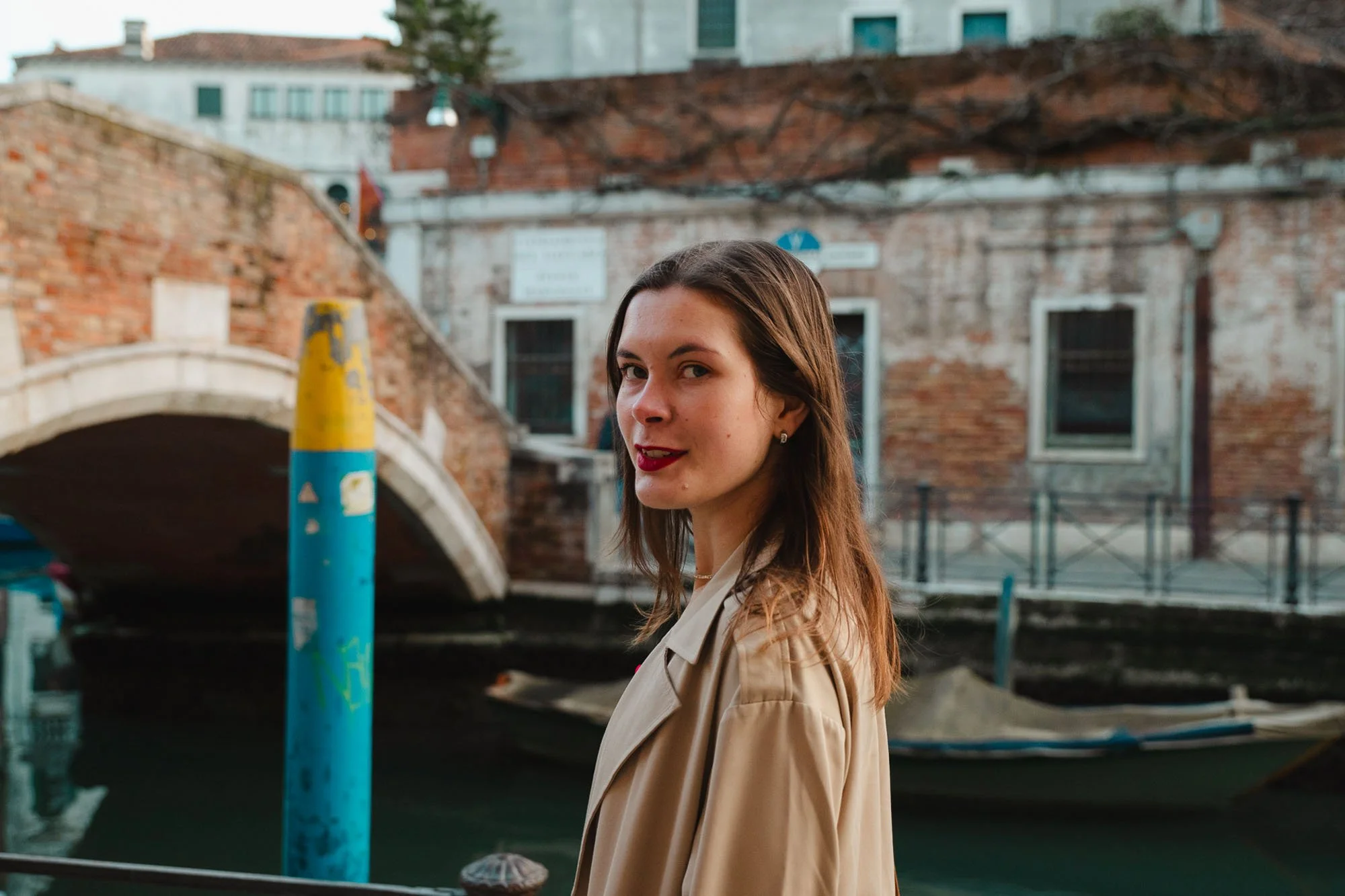 Ragazza con capelli lunghi e rossetto rosso che si trova vicino a un canale a Venezia, con un ponte di pietra e edifici antichi sullo sfondo.
