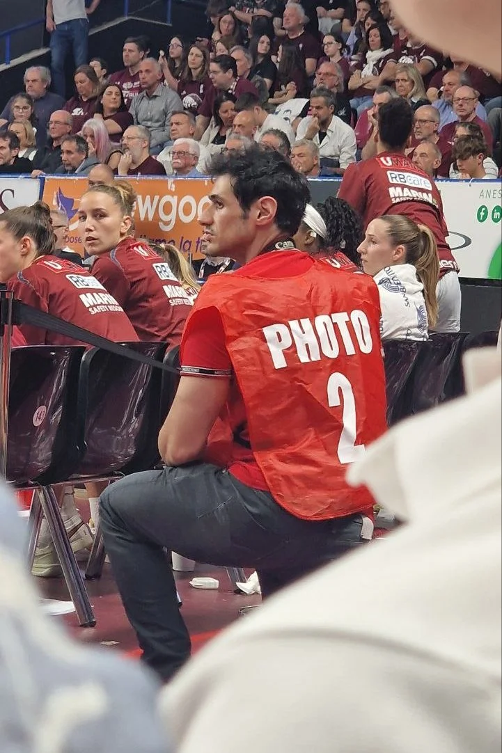 Un uomo seduto con una maglietta rossa con un cartellino che dice 'PHOTO 2' sulla schiena, circondato da altre persone in un evento pubblico o sportivo, con una folla seduta sullo sfondo.