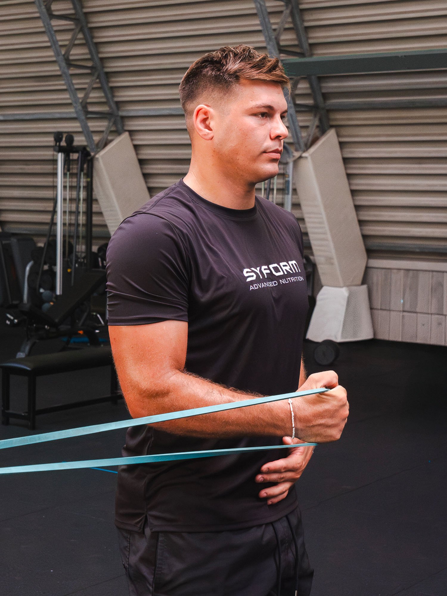 Uomo giovane con capelli corti e chiari, vestito con una maglietta nera con scritto 'SY-ORM Advanced Nutrition', che fa esercizi con una torre di sbarra per trazioni in un ambiente di palestra.
