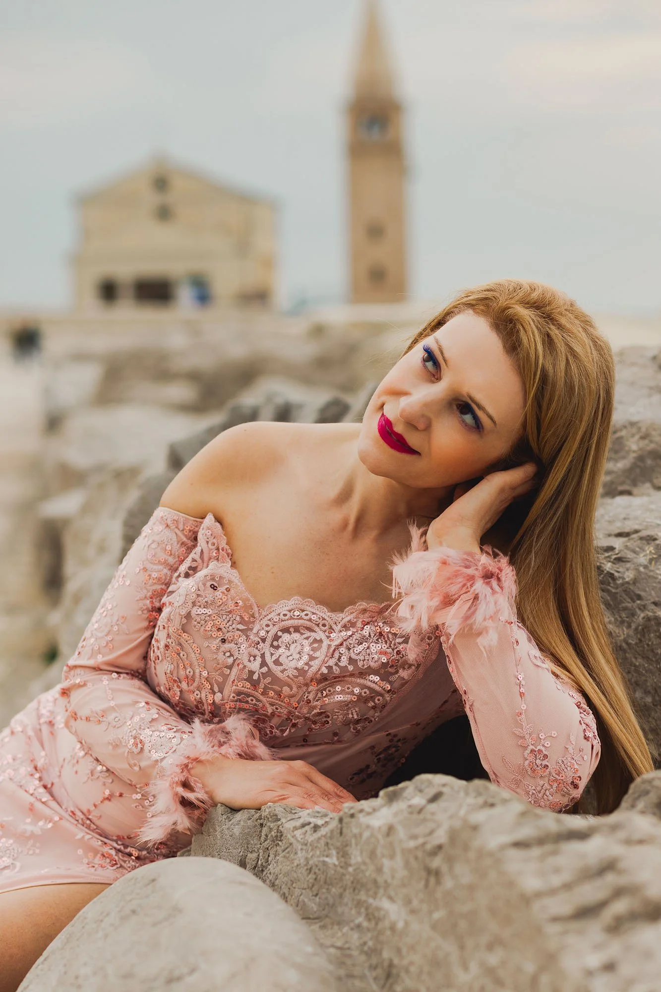 Un ritratto di una donna con capelli rossi, vestita con un abito rosa con ricami e dettagli in pelliccia rosa, che si sdraia su rocce vicino al mare, con uno sfondo di un edificio storico con torre.