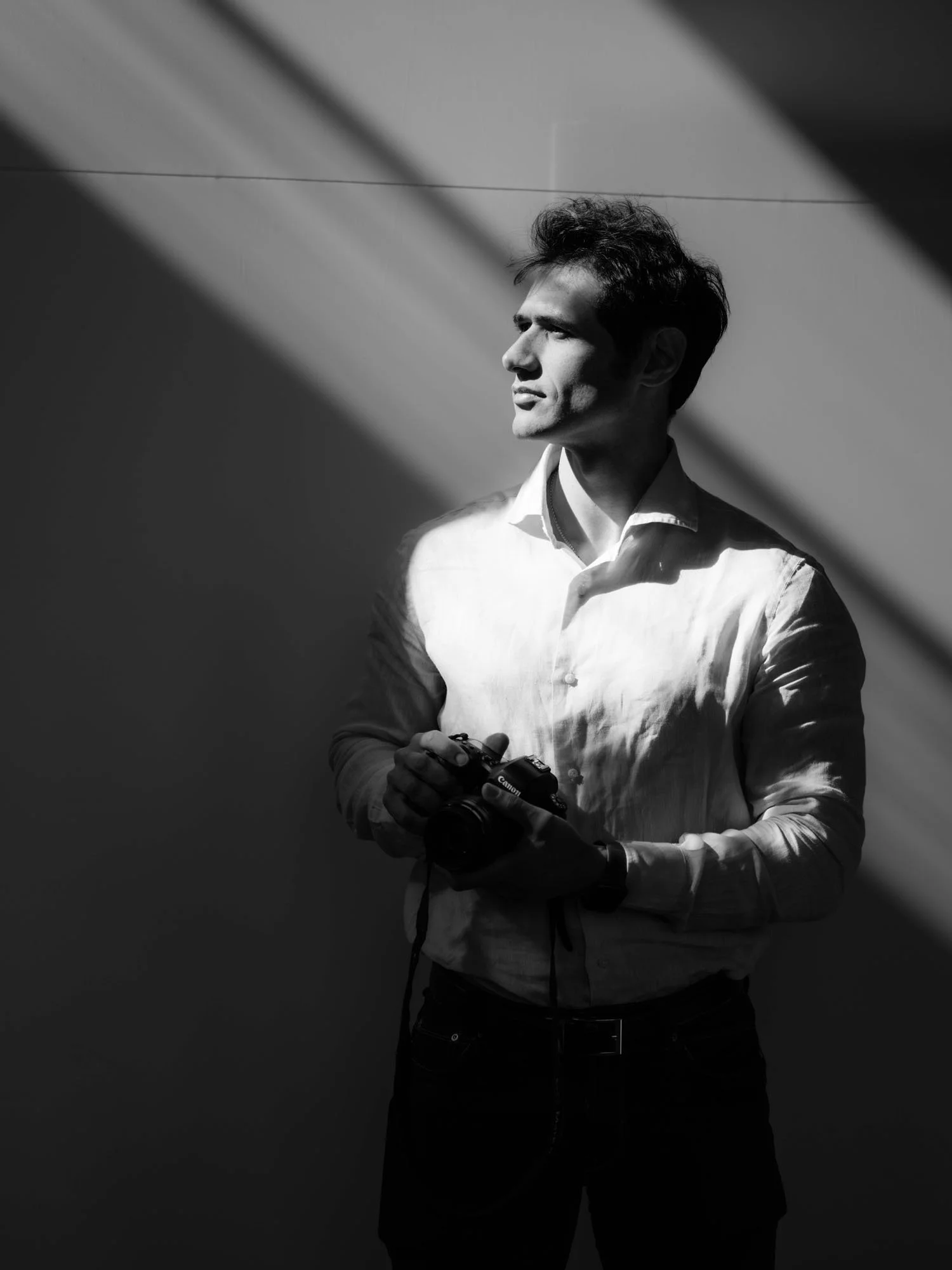 Uomo con camicia bianca e capelli ricci, con una telecamera in mano, che guarda verso l'alto, in controluce su uno sfondo chiaro e forme di luce e ombra.