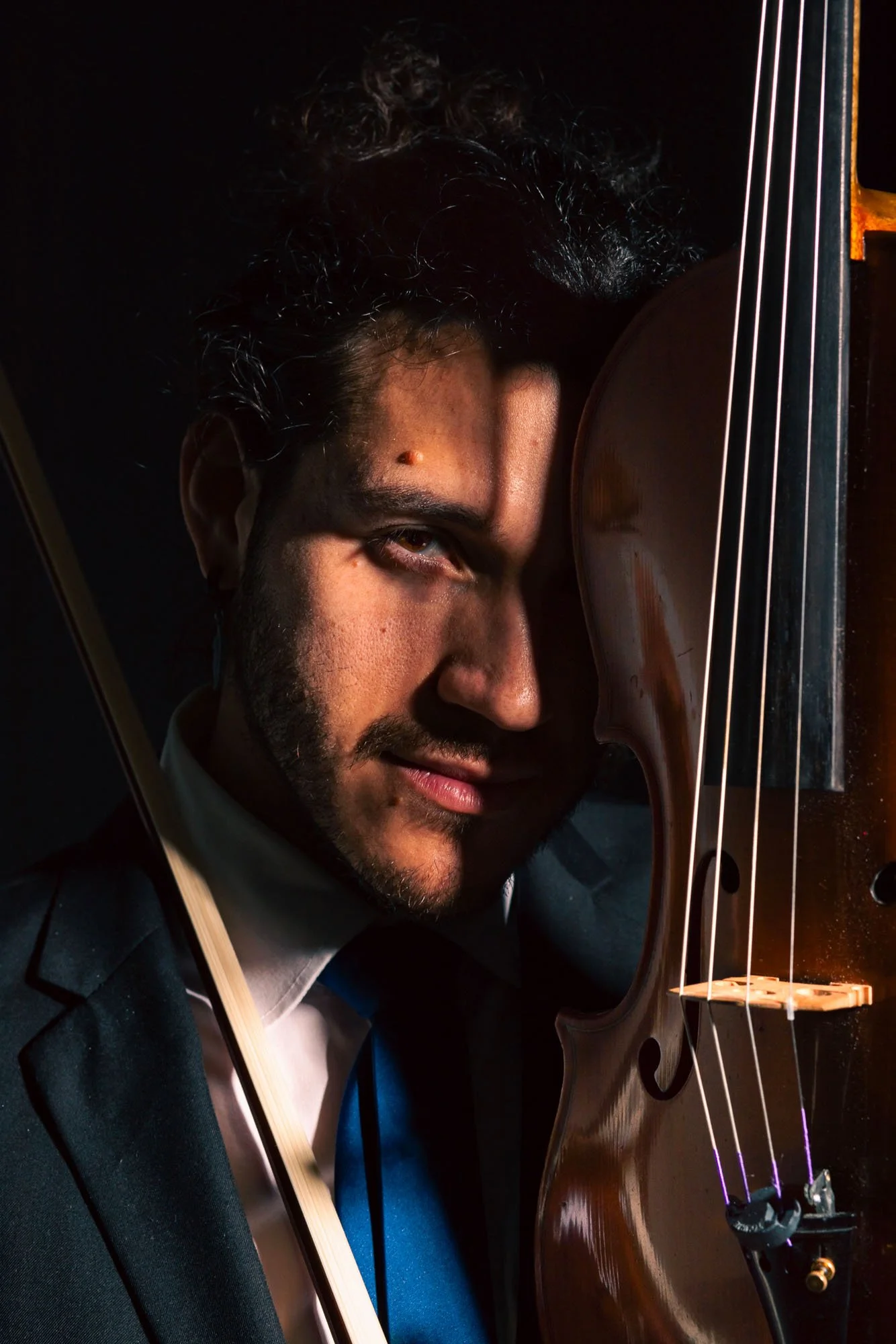 Uomo con barba e capelli ricci si trova vicino a un violino e un arco, in abito elegante, con studio di musica domestico in sfondo.