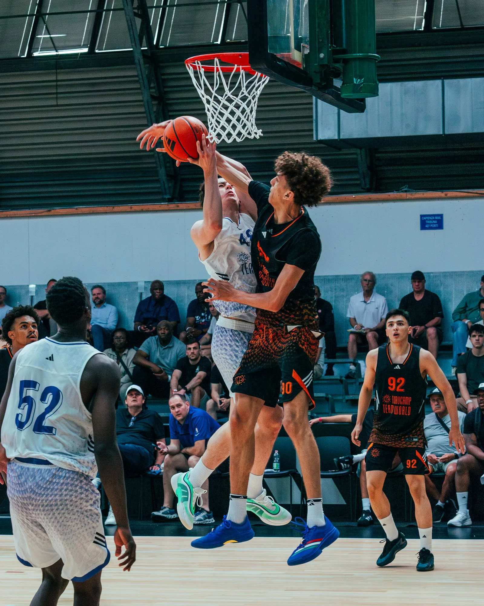 Giocatori di basket in azione durante una partita, con pubblico che guarda sullo sfondo.