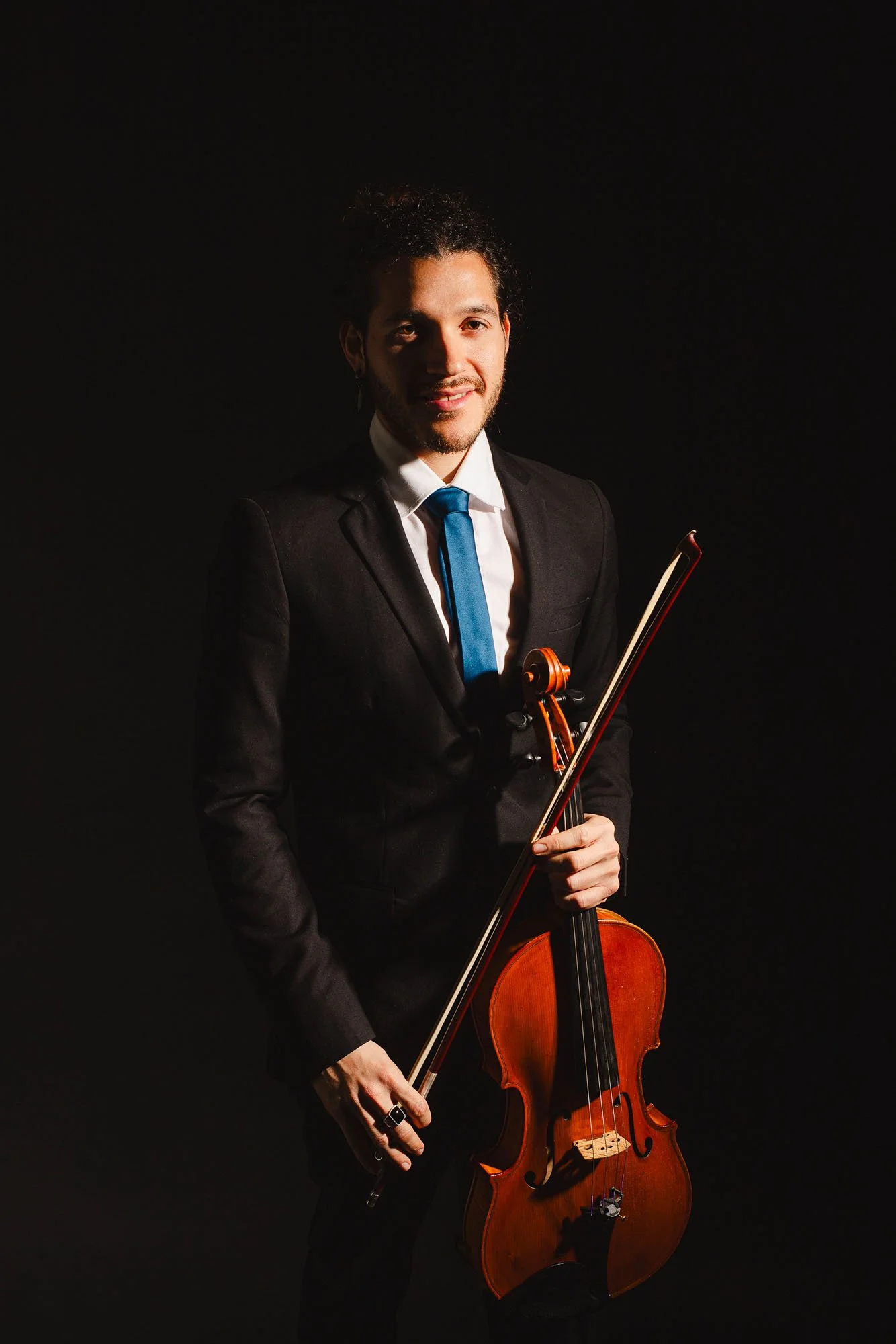 Uomo vestito in abito nero con cravatta blu, tiene un violino e un arco, sfondo nero, luce focalizzata sul volto e sul violino.