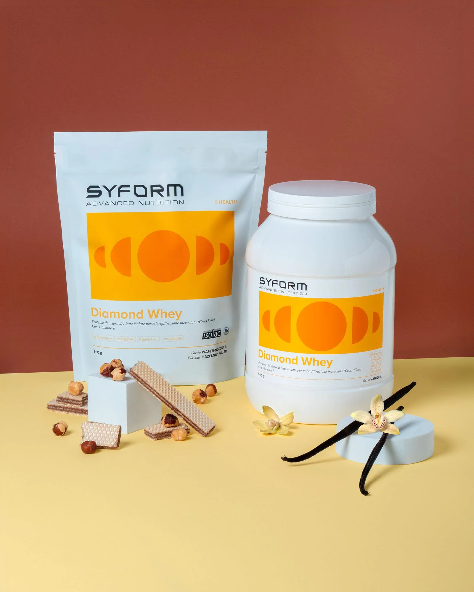 Due scatole di integratori di proteine Syform Diamond Whey, una in polvere e l'altra in barattolo, con merendine, nocciole, panna e fiori bianchi decorativi, su uno sfondo giallo.