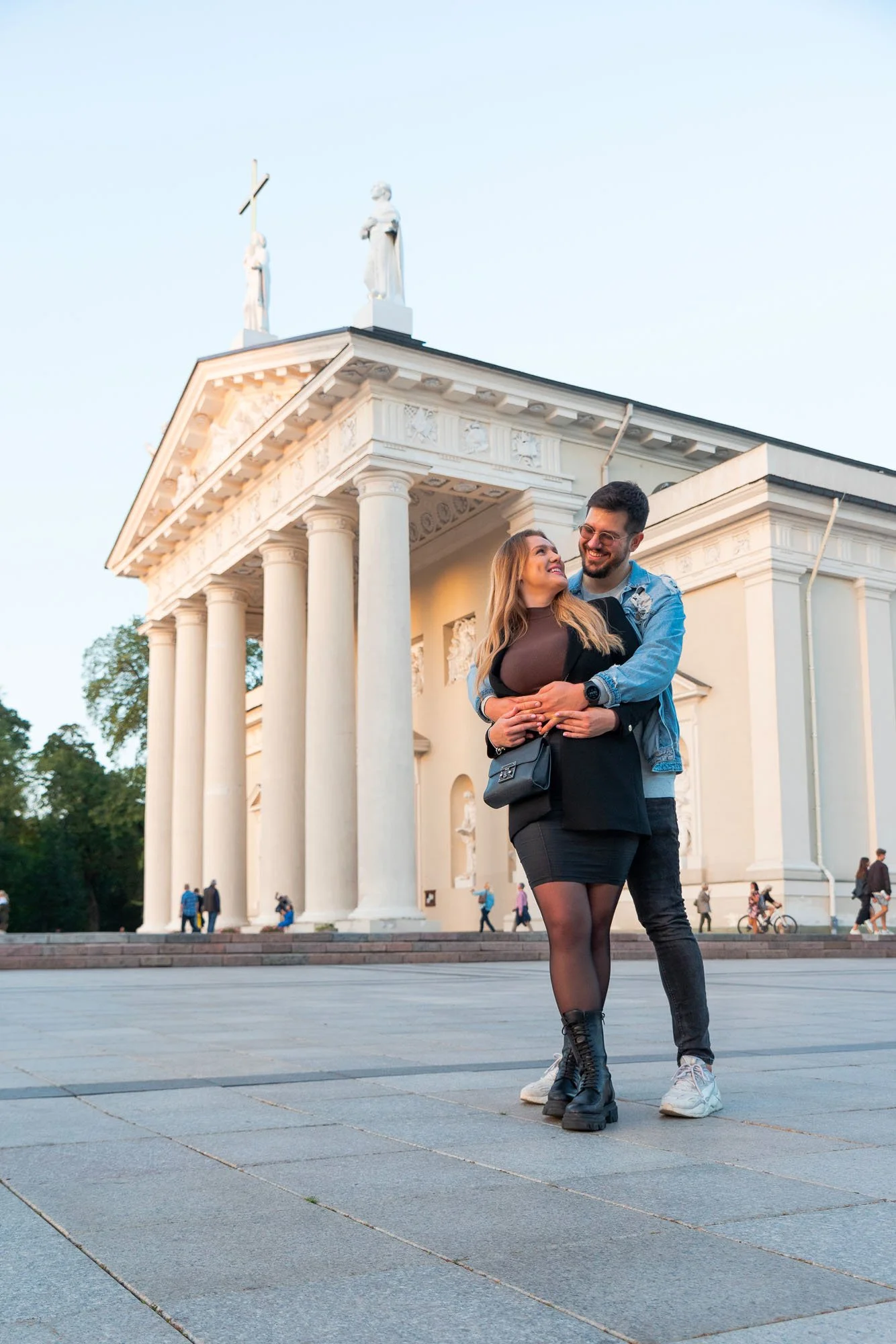 Una coppia di giovani si abbraccia e sorride di fronte a una grande chiesa con colonne e statue sul tetto, al tramonto.